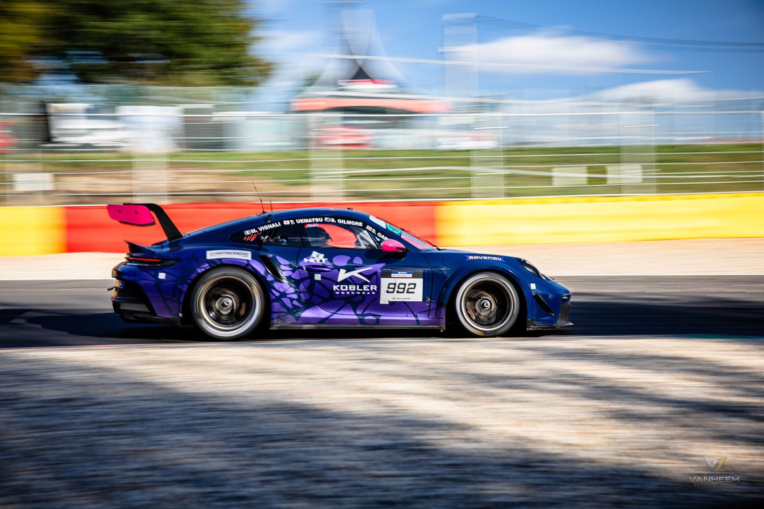 A blue and purple Porsche GT3 race car speeds around a track circuit during a motorsport racing event.