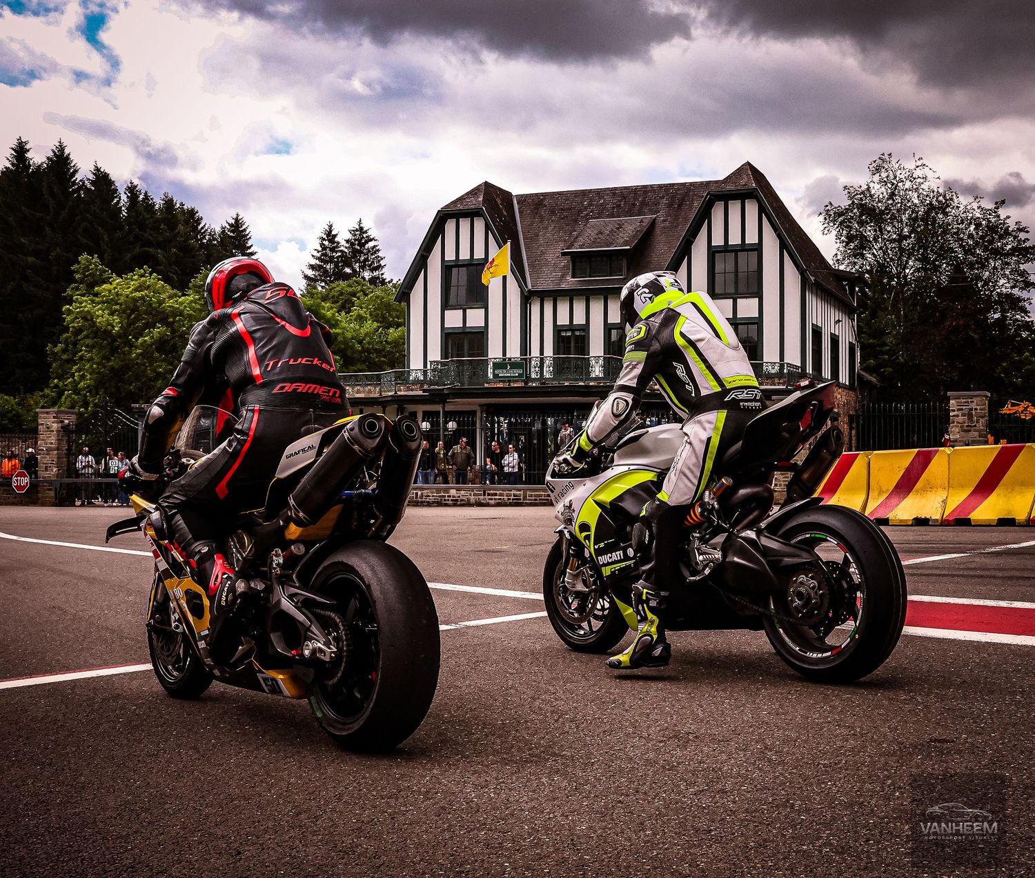 Two motorcycle racers on sport bikes prepare to race on a wet circuit track with a Tudor-style building in the background.
