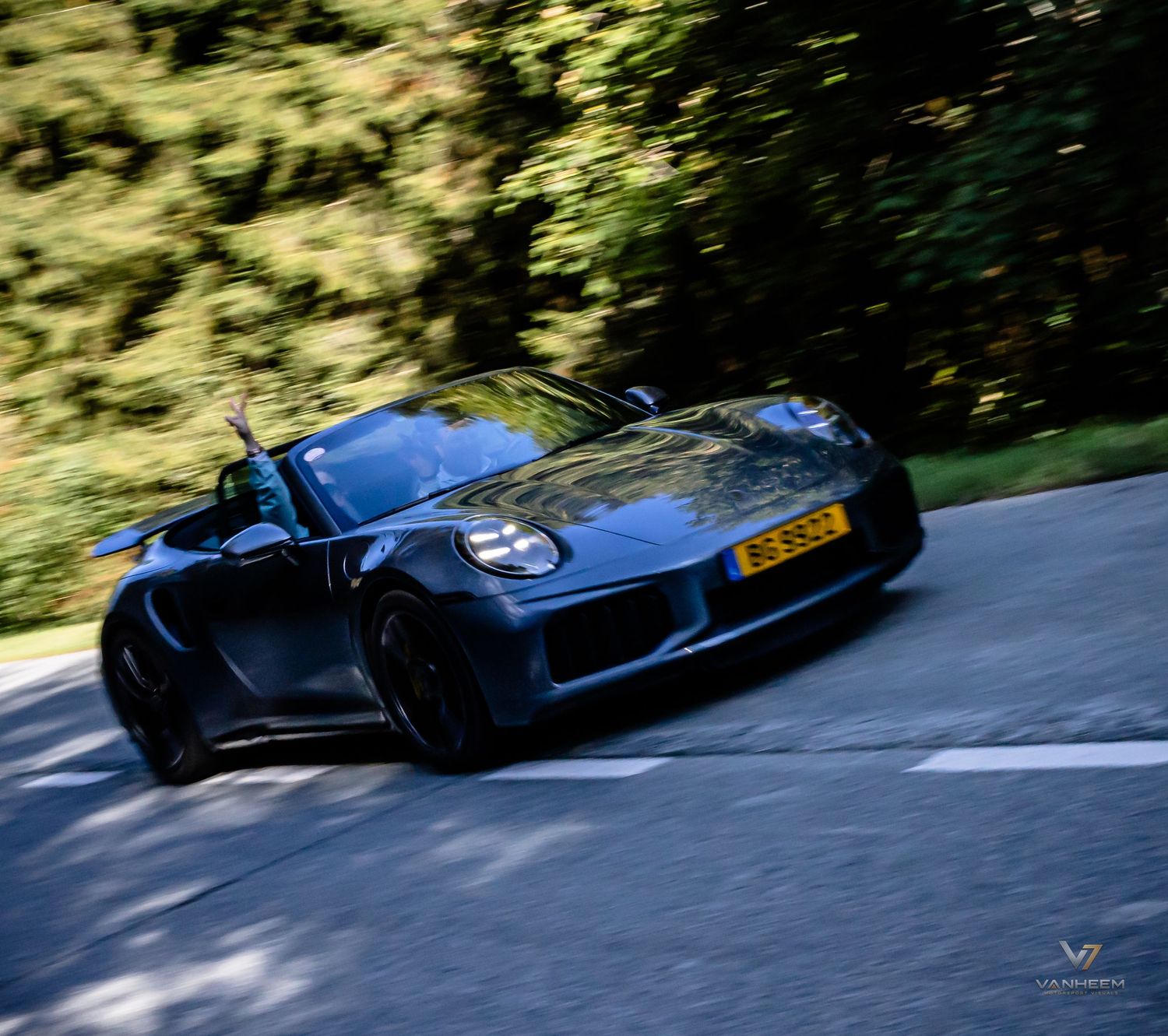 Dark blue Porsche 911 Speedster convertible driving on a winding road surrounded by lush green trees.