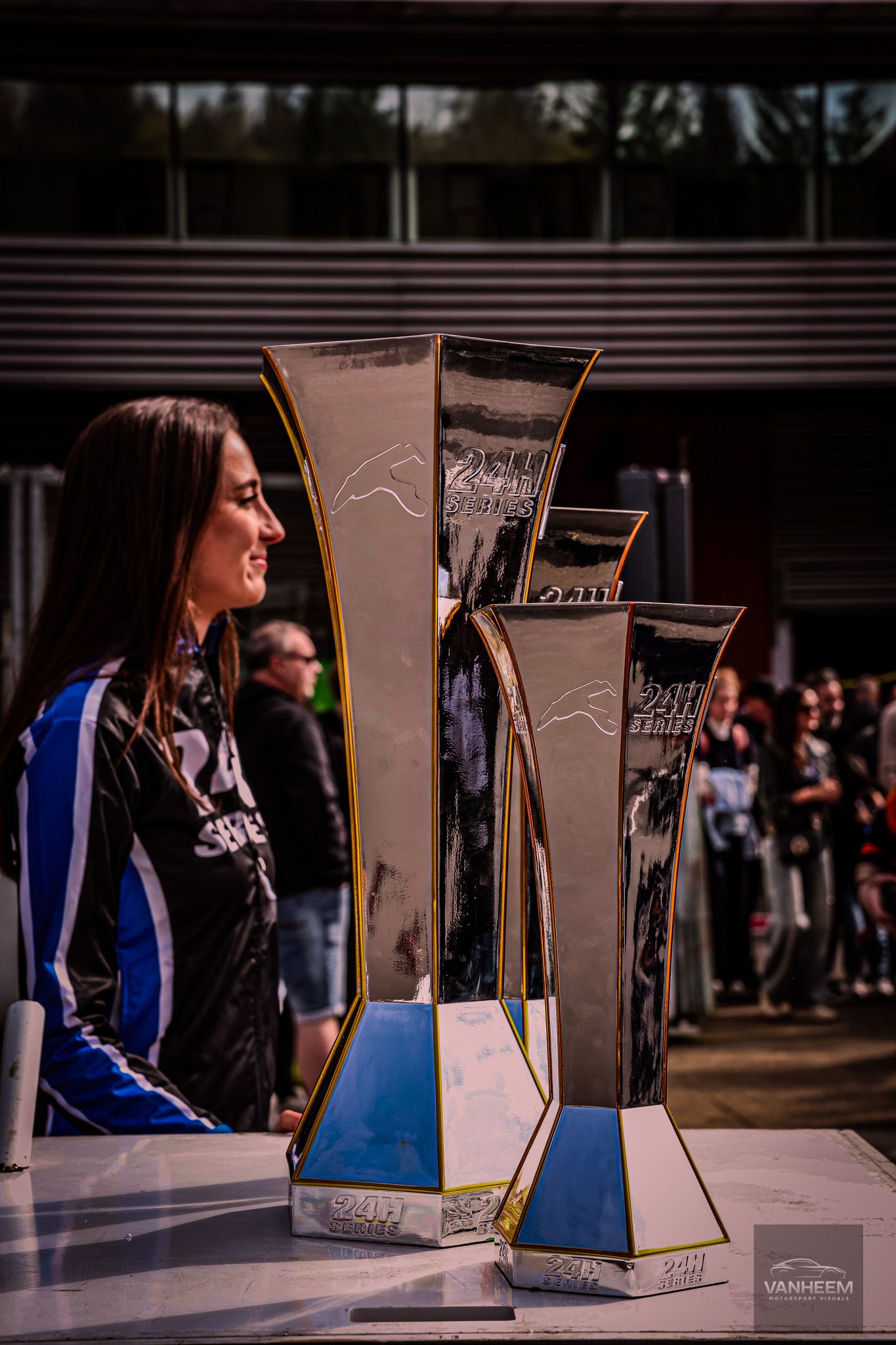 Woman admiring large shiny racing trophies displayed outdoors at a motorsport awards ceremony.