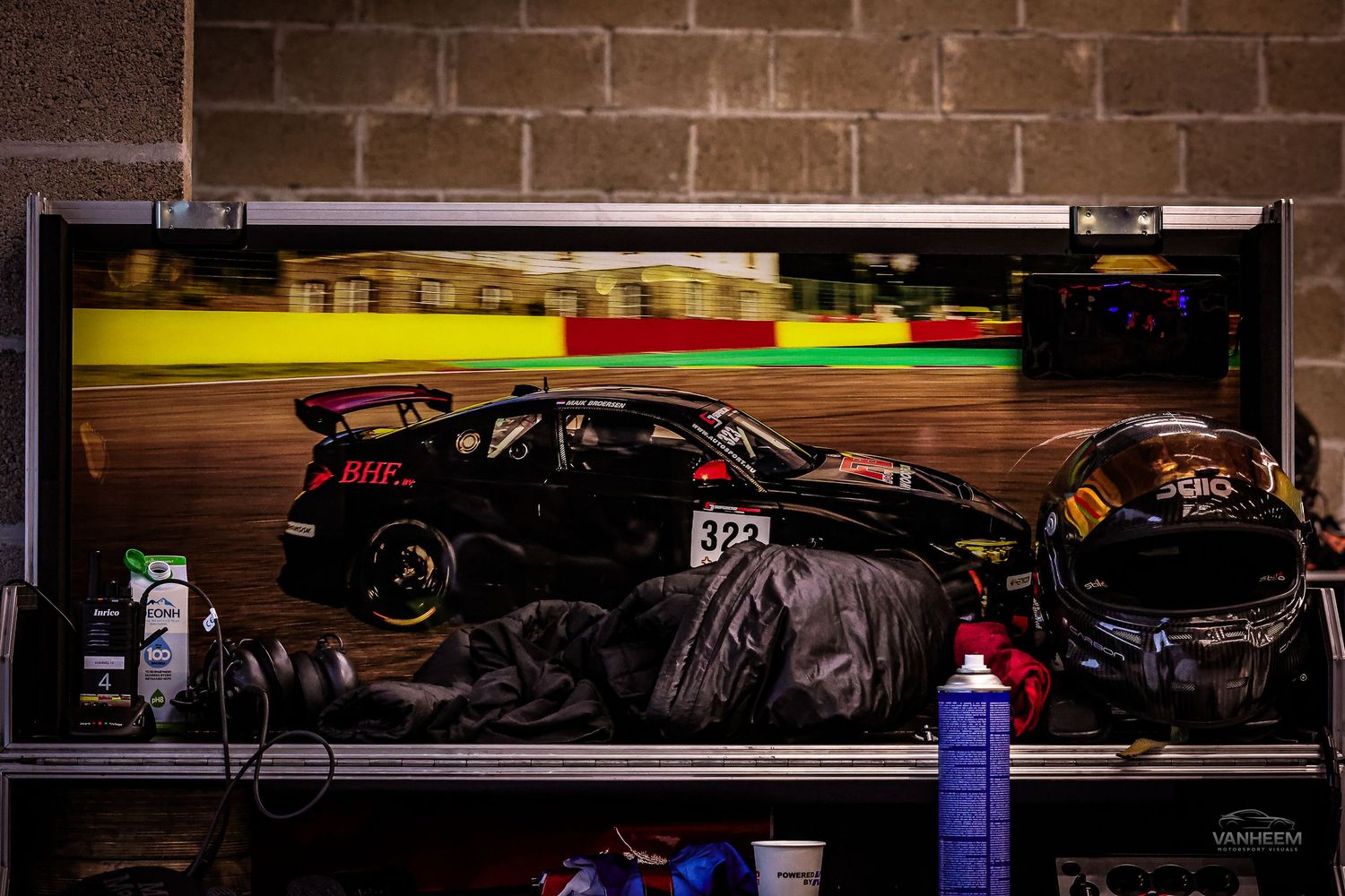 Racing car number 321 displayed on a monitor in a pit garage with helmets and equipment on a workbench.