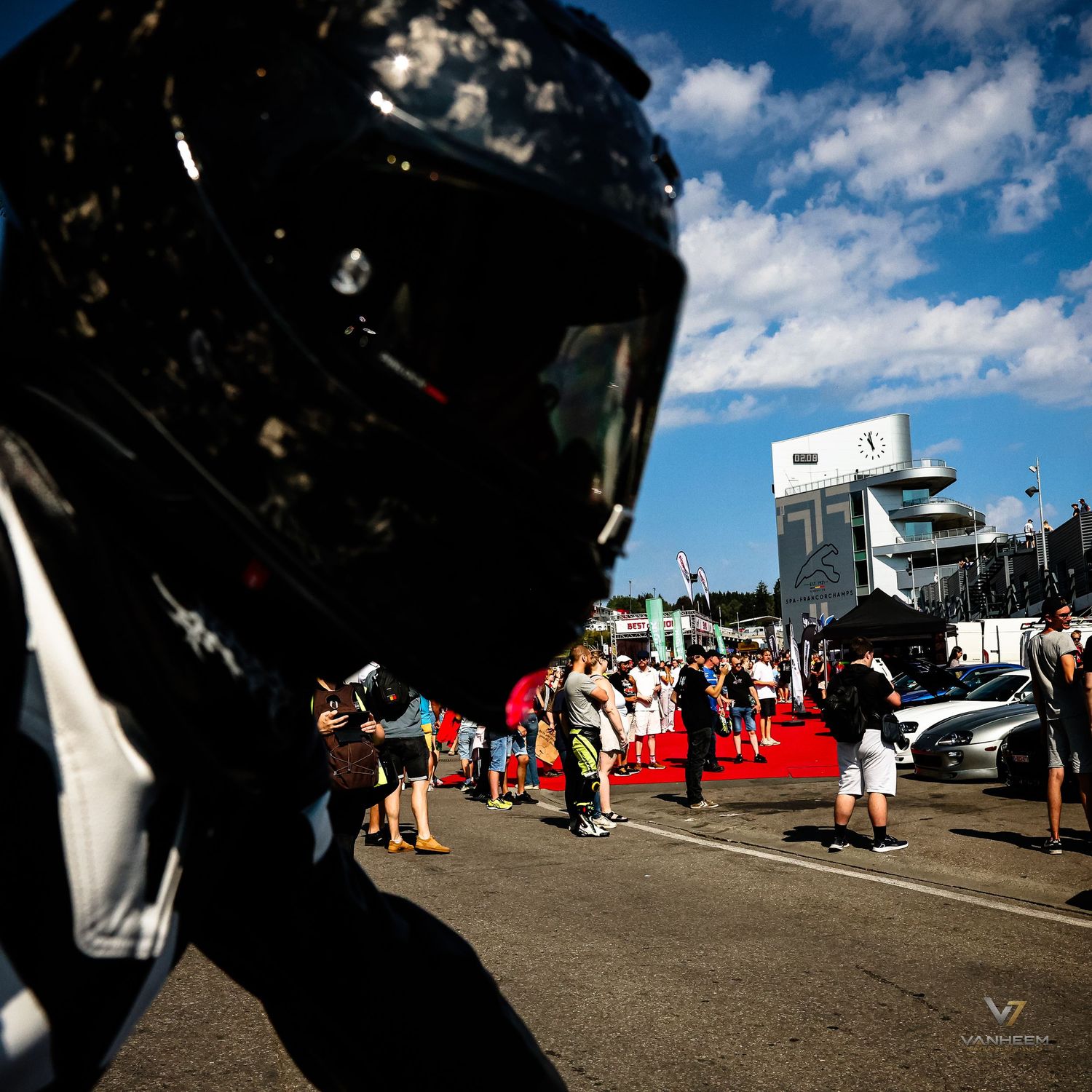 A racing helmet in the foreground at a motorsport event with spectators and pit lane visible under blue skies.