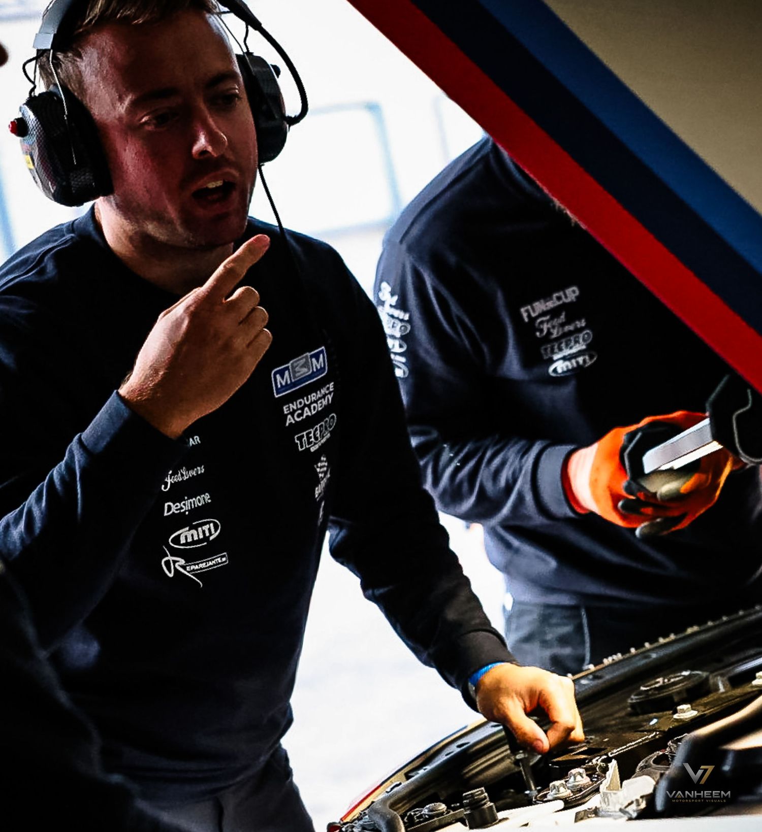 Two mechanics in branded racing team uniforms work on a car engine in a professional motorsport pit garage.
