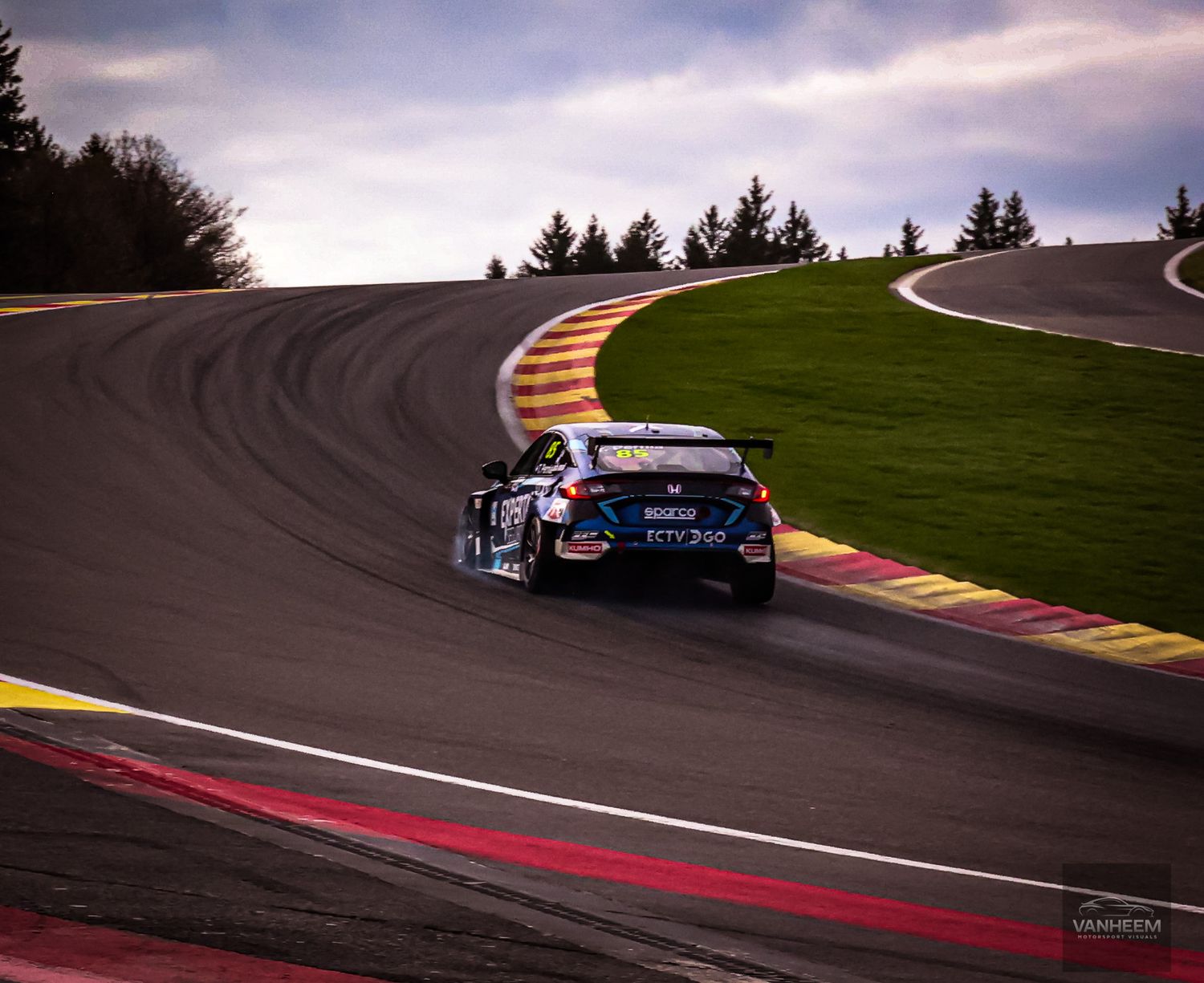 A race car navigates a sweeping curve at Spa-Francorchamps circuit under dramatic cloudy skies.