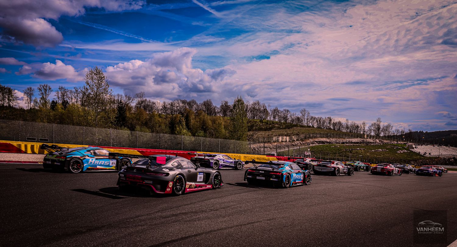 Multiple GT racing cars lined up on a race track circuit under a blue sky with clouds and green trees in background.