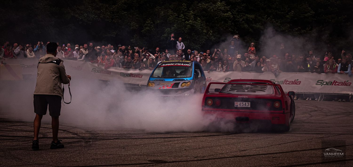 Two cars drifting on a track surrounded by smoke and spectators at an outdoor motorsport event.