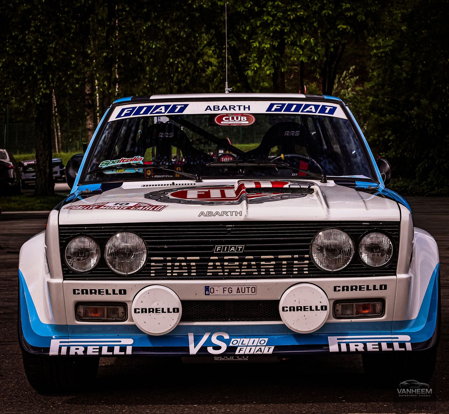 A Fiat Abarth rally car with blue and white livery, Pirelli tires, and Carello lights on display at an outdoor event.
