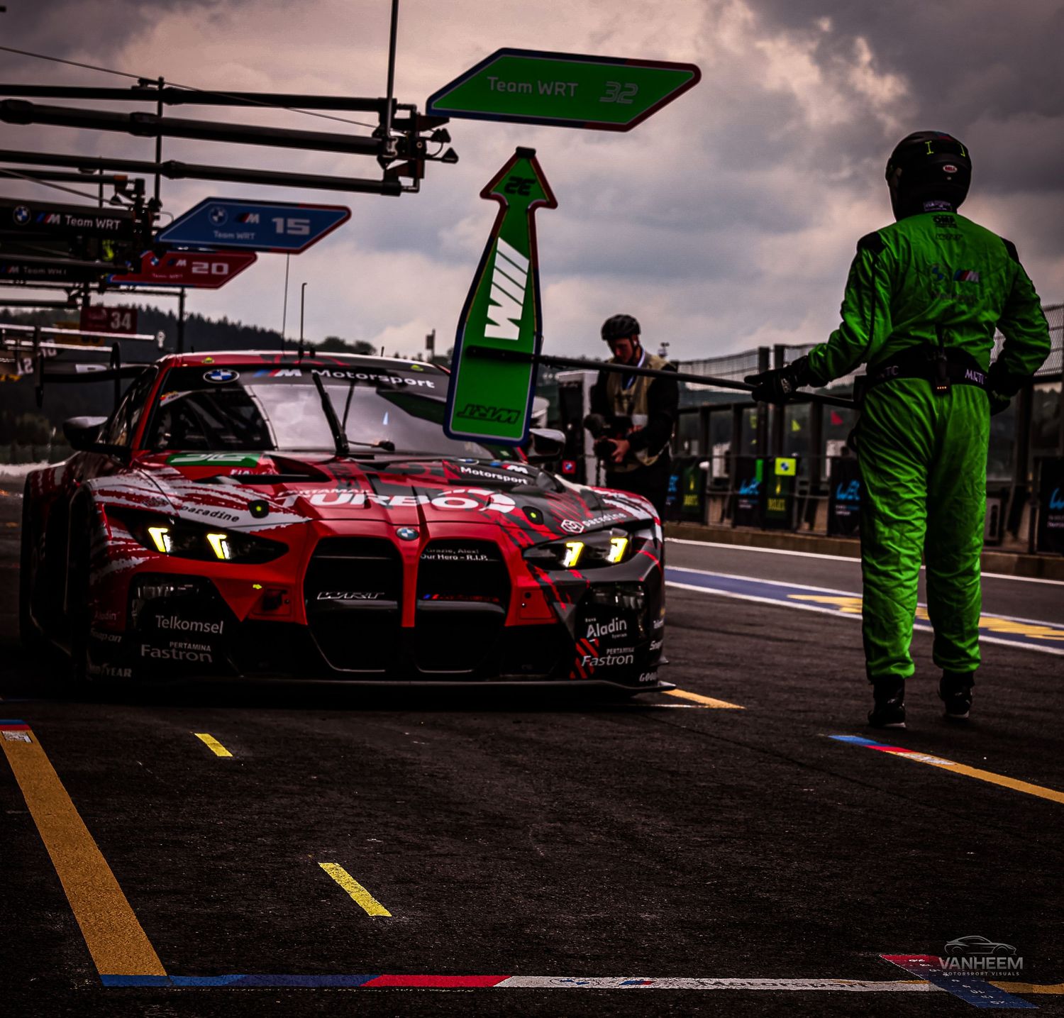 A red Toyota GR racing car exits pit lane at a motorsport event, with a crew member in green suit standing nearby.