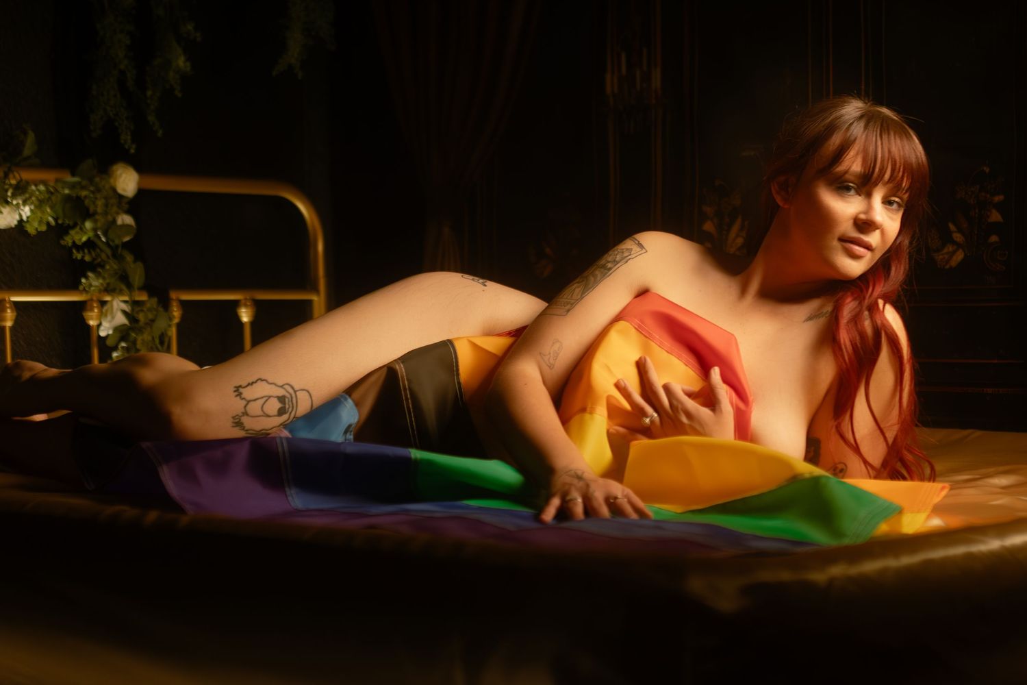 Person draped in Progress Pride flag on a golden bed during inclusive boudoir session at Lock & Key Photography in Fort Worth, TX.