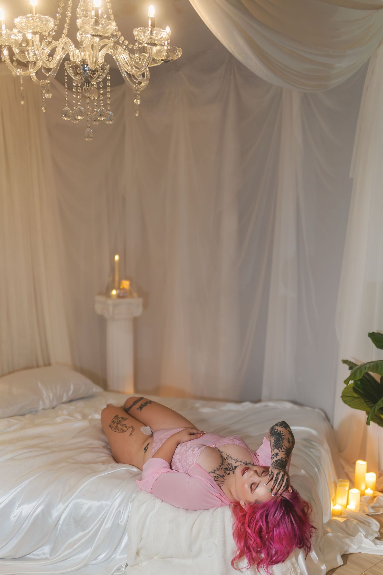 Woman with vivid pink hair lying on satin in candlelit Fort Worth boudoir studio with chandelier overhead.