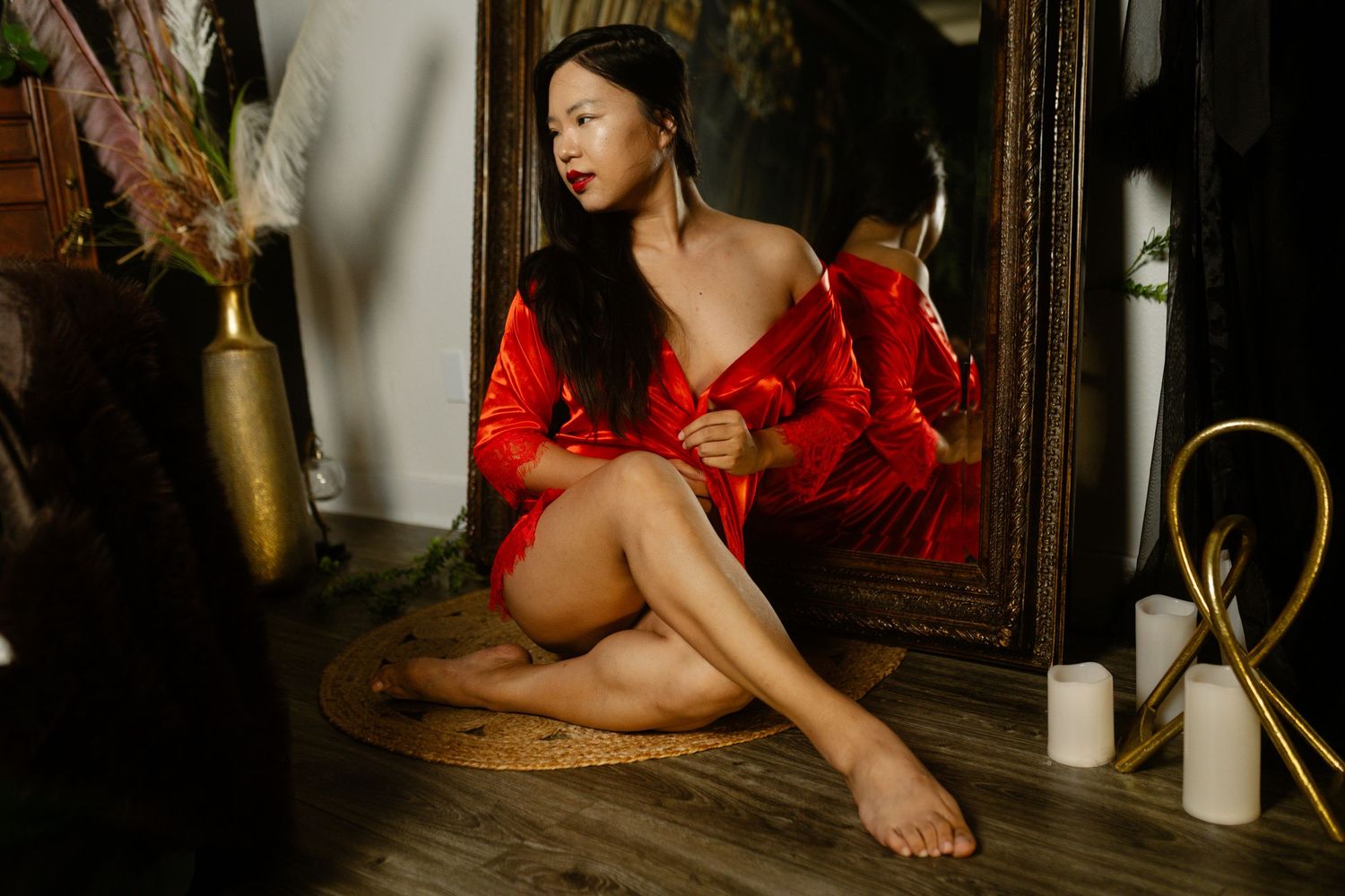 DFW boudoir photographer- woman in red satin lingerie posed by mirror with warm candlelight and elegant decor in this boudoir photo.