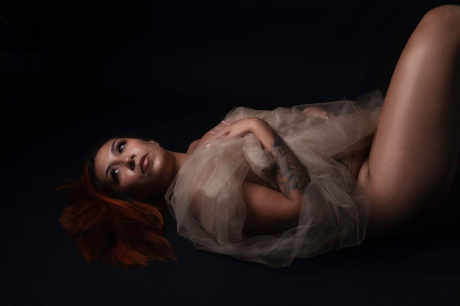 Tattooed woman wrapped in sheer tulle against black backdrop, DFW boudoir photography by Lock & Key Fort Worth.