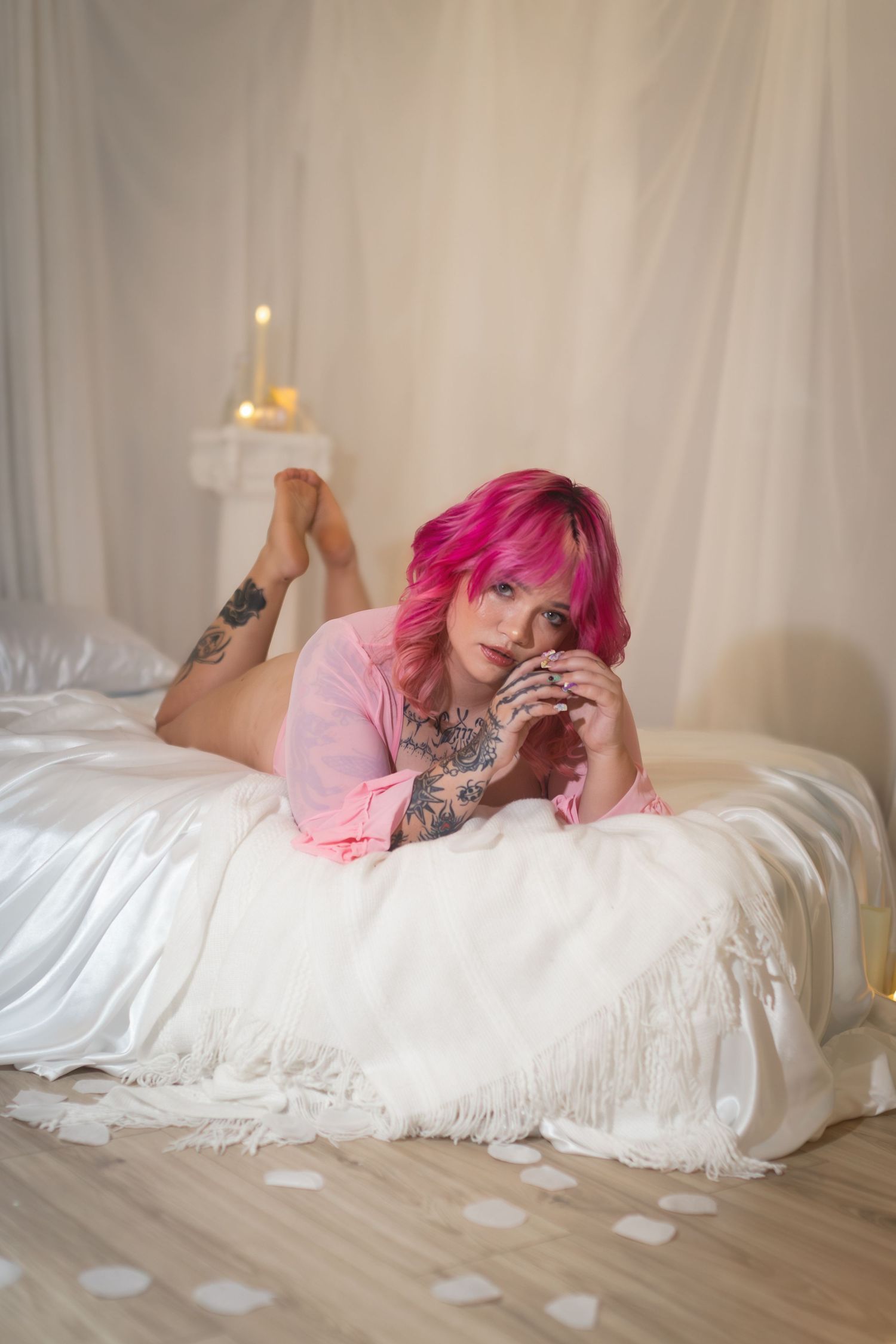 Pink-haired tattooed woman lying on white bed in pink robe, romantic candlelit boudoir portrait.