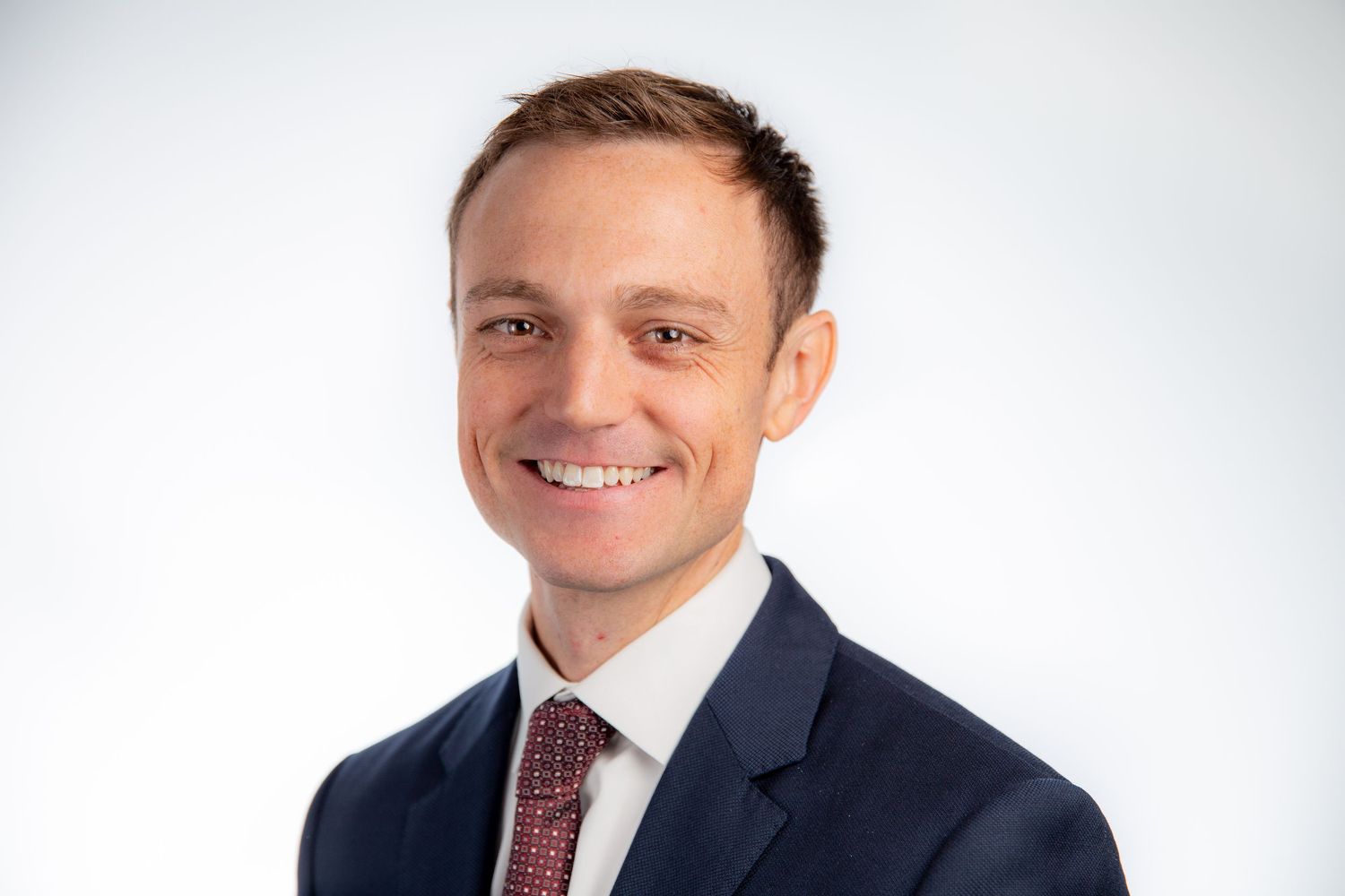 Smiling professional man in navy suit and red tie against white background headshot.