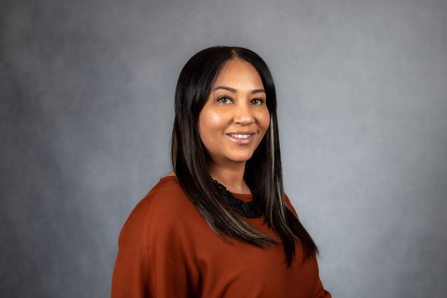 Professional headshot of a smiling woman with long dark hair wearing a rust-colored sweater against a gray background.