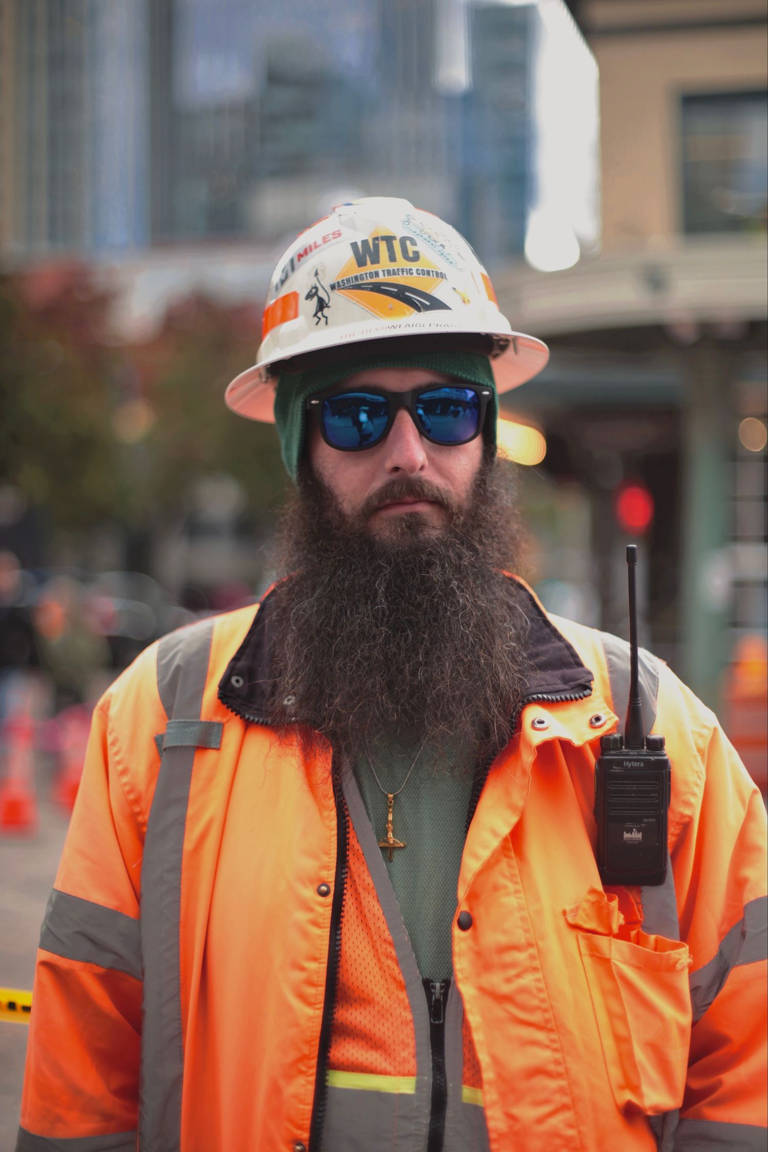 Bearded construction worker in orange safety jacket and WTC hard hat holds radio on city street.