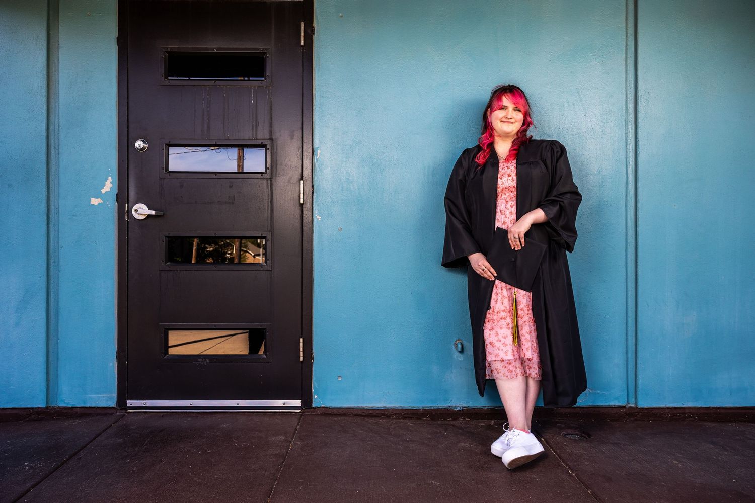 Woman with pink hair in black coat leaning against teal wall beside a dark door.