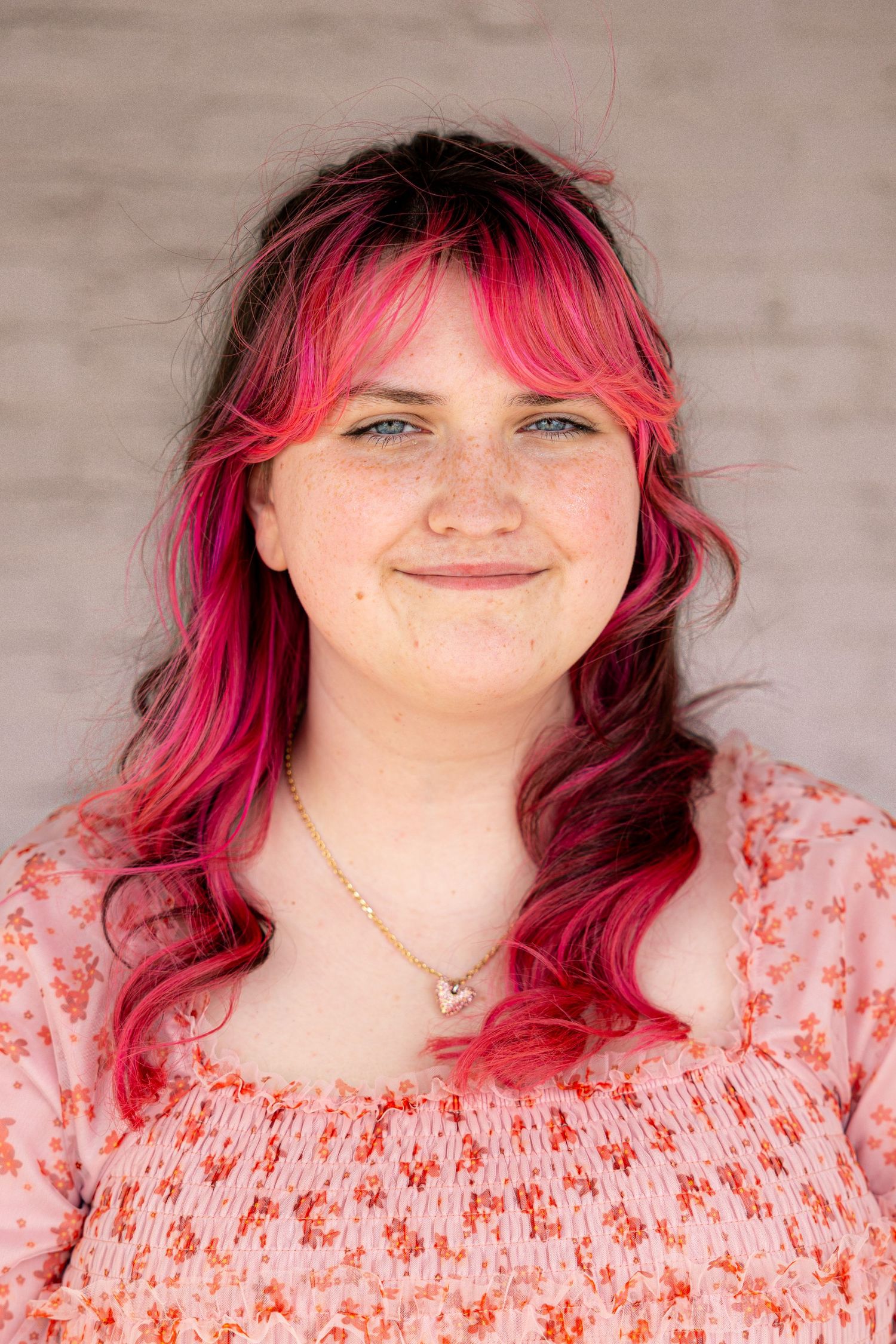 Young woman with vibrant pink and red dyed hair, freckles, and a floral top smiling against a white brick wall.