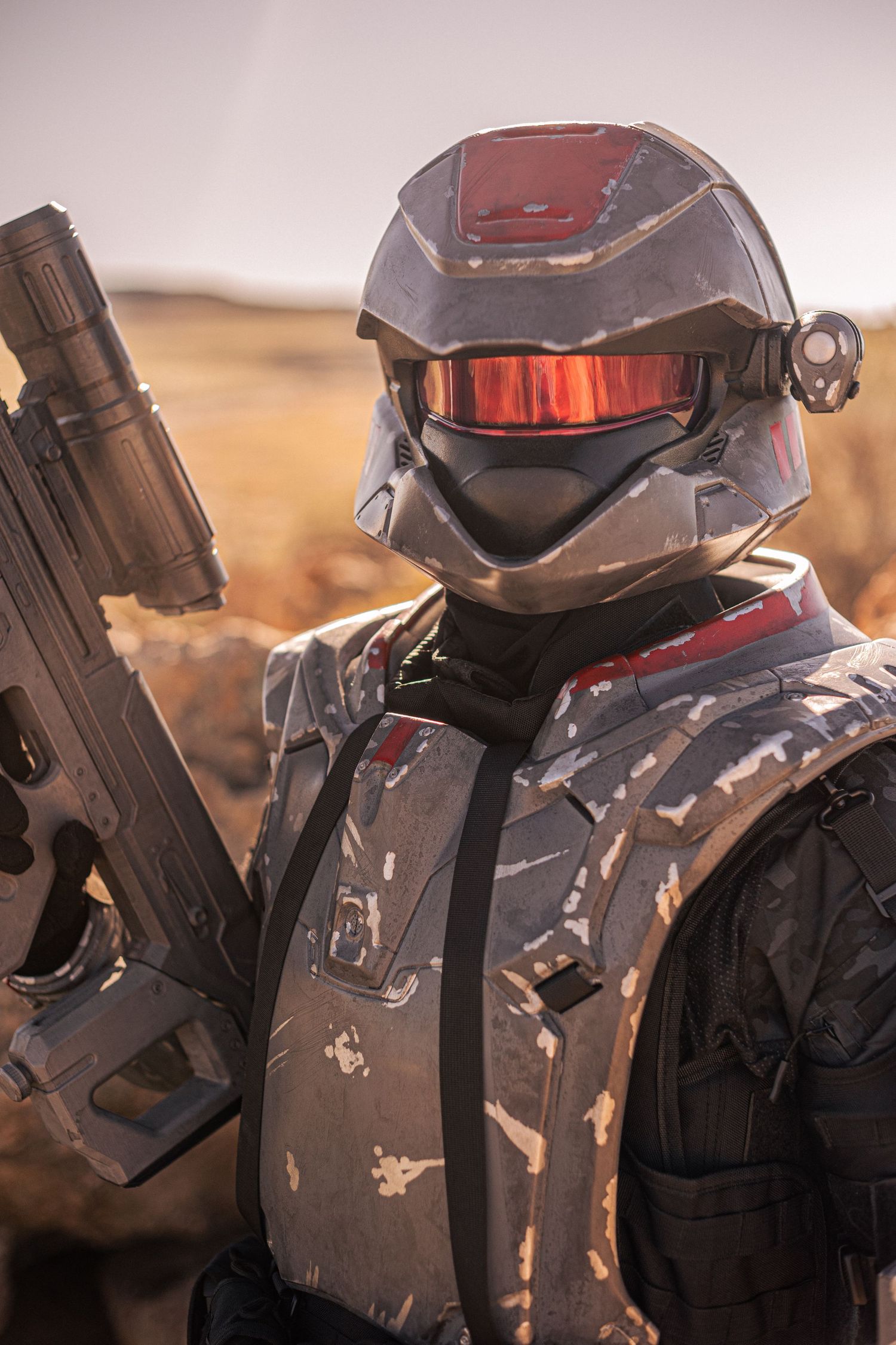 Close-up of weathered sci-fi soldier helmet with red visor holding large weapon in desert.