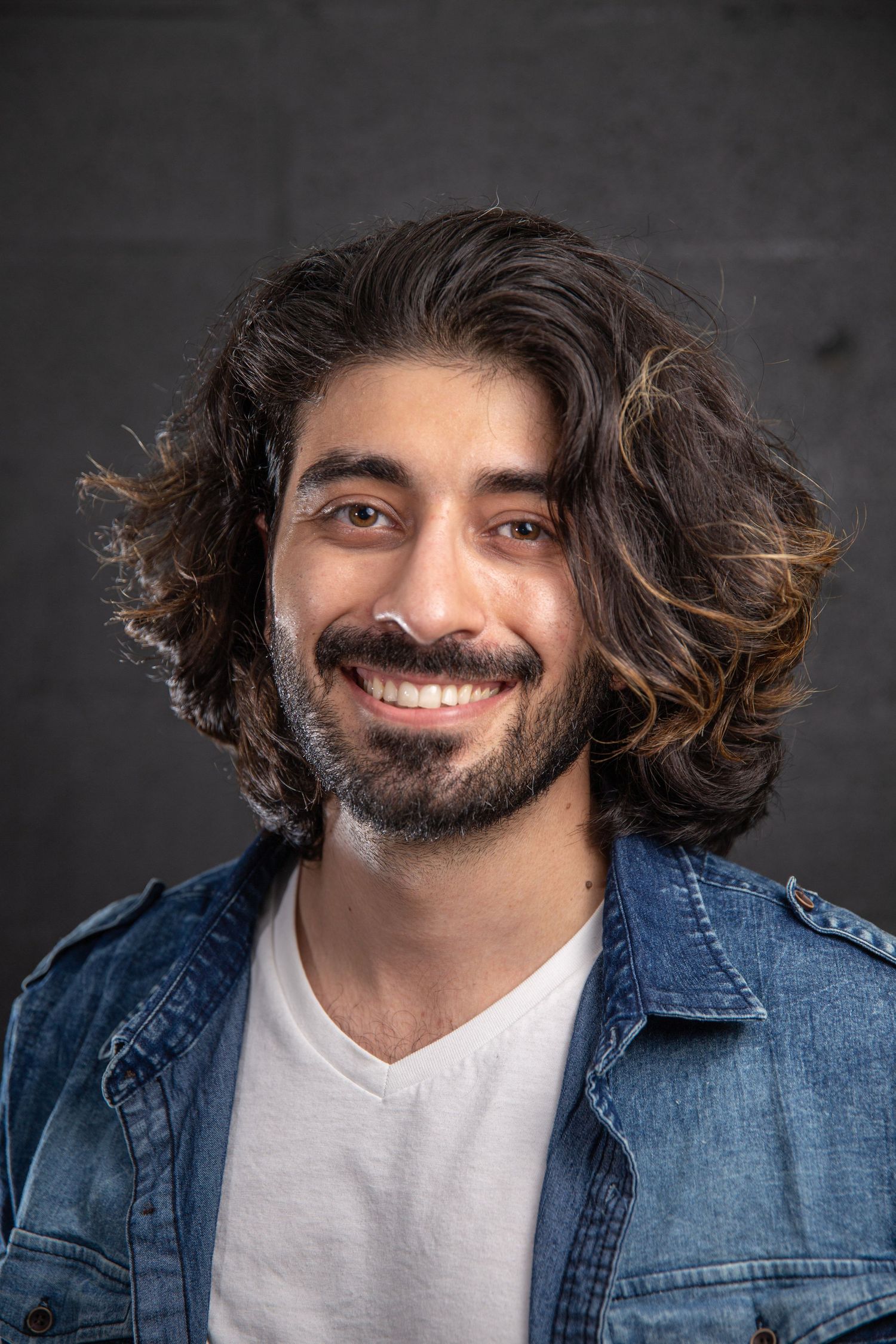 Young man with long curly brown hair and beard wearing a denim jacket, smiling headshot on dark background.