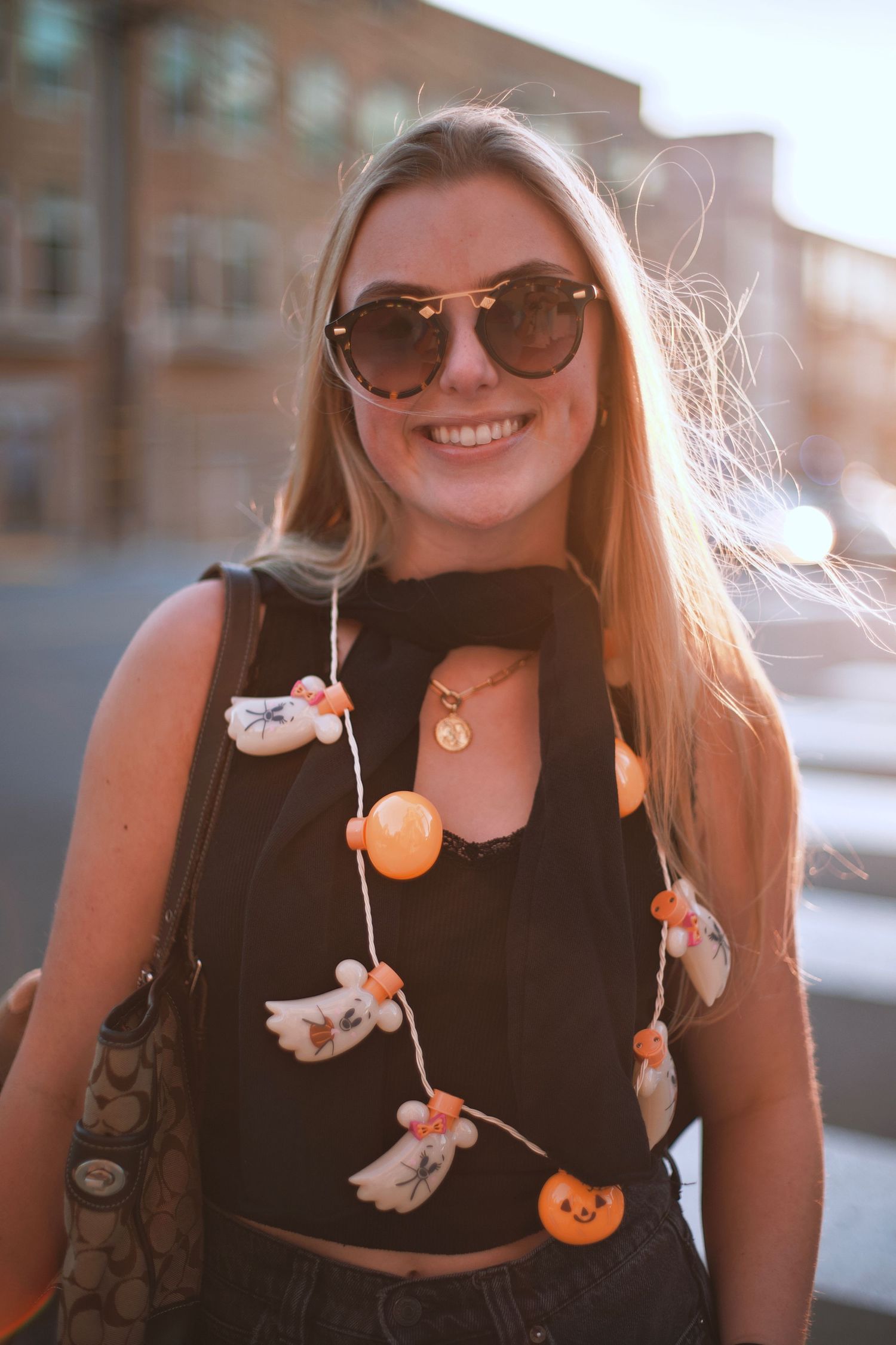 Smiling blonde woman wearing sunglasses and Halloween garland necklace outdoors in golden hour light.