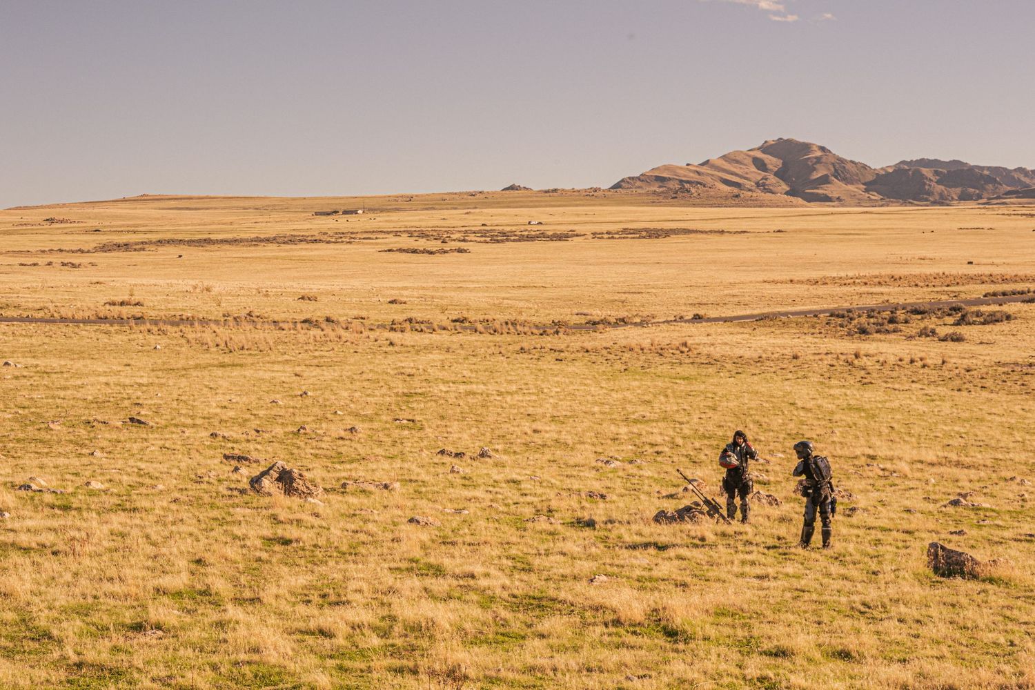Two people in tactical gear stand in a vast golden grassland with a rocky mountain in the background.