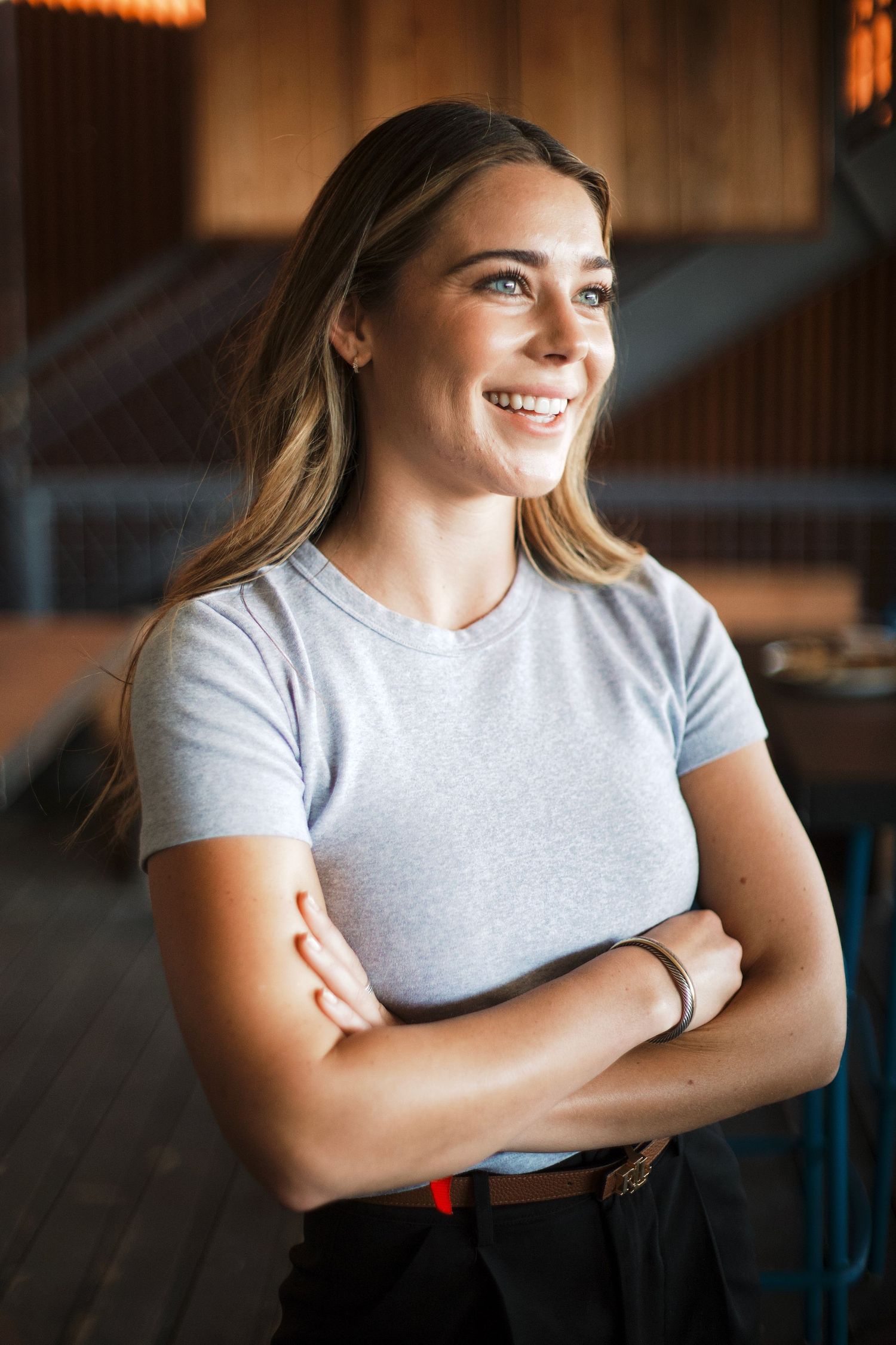 Smiling young woman with long blonde hair in gray t-shirt standing with arms crossed indoors.