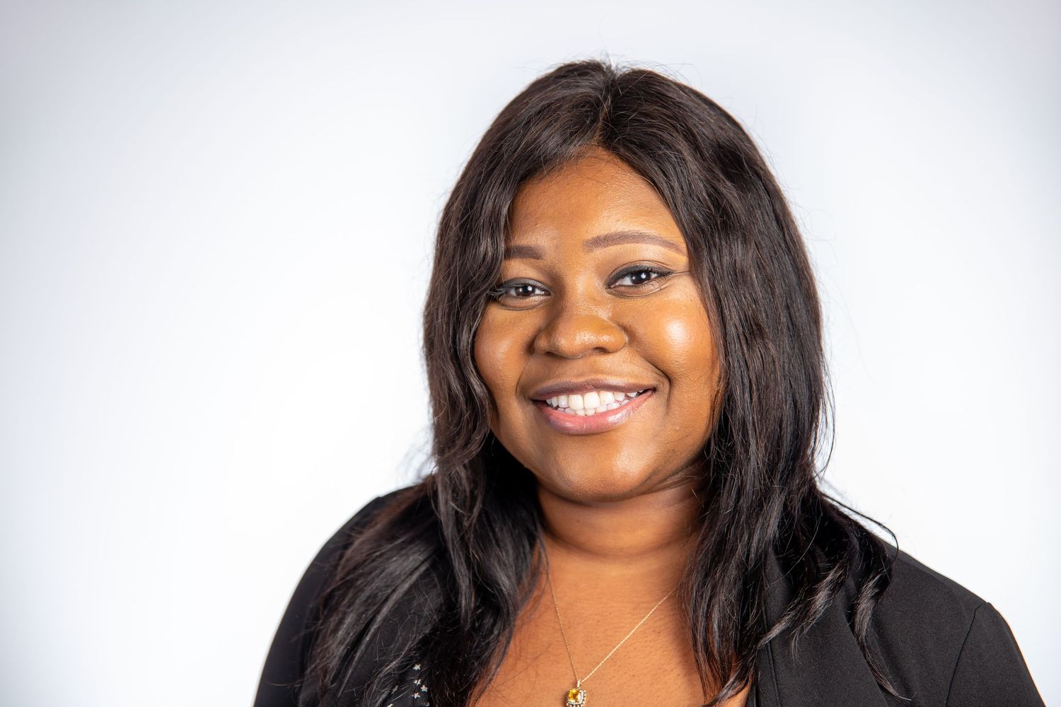 Smiling woman with long dark hair wearing black blazer and gold necklace on white background.