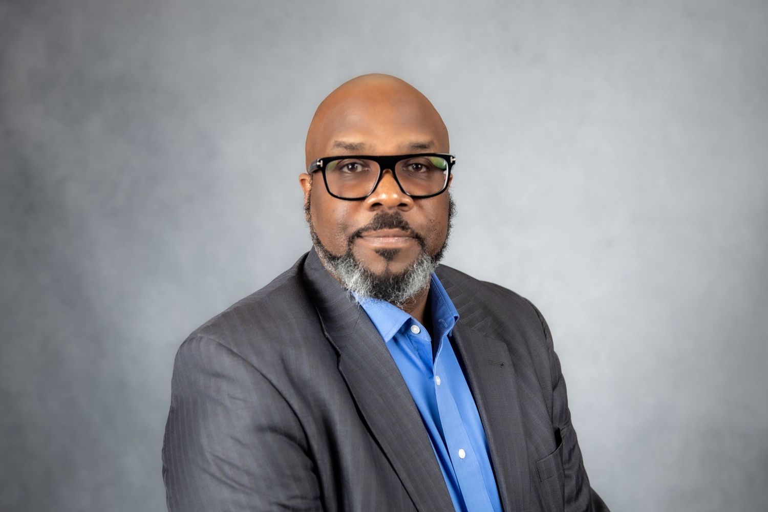 Professional headshot of a bald man with glasses and a beard wearing a blue shirt and gray blazer.