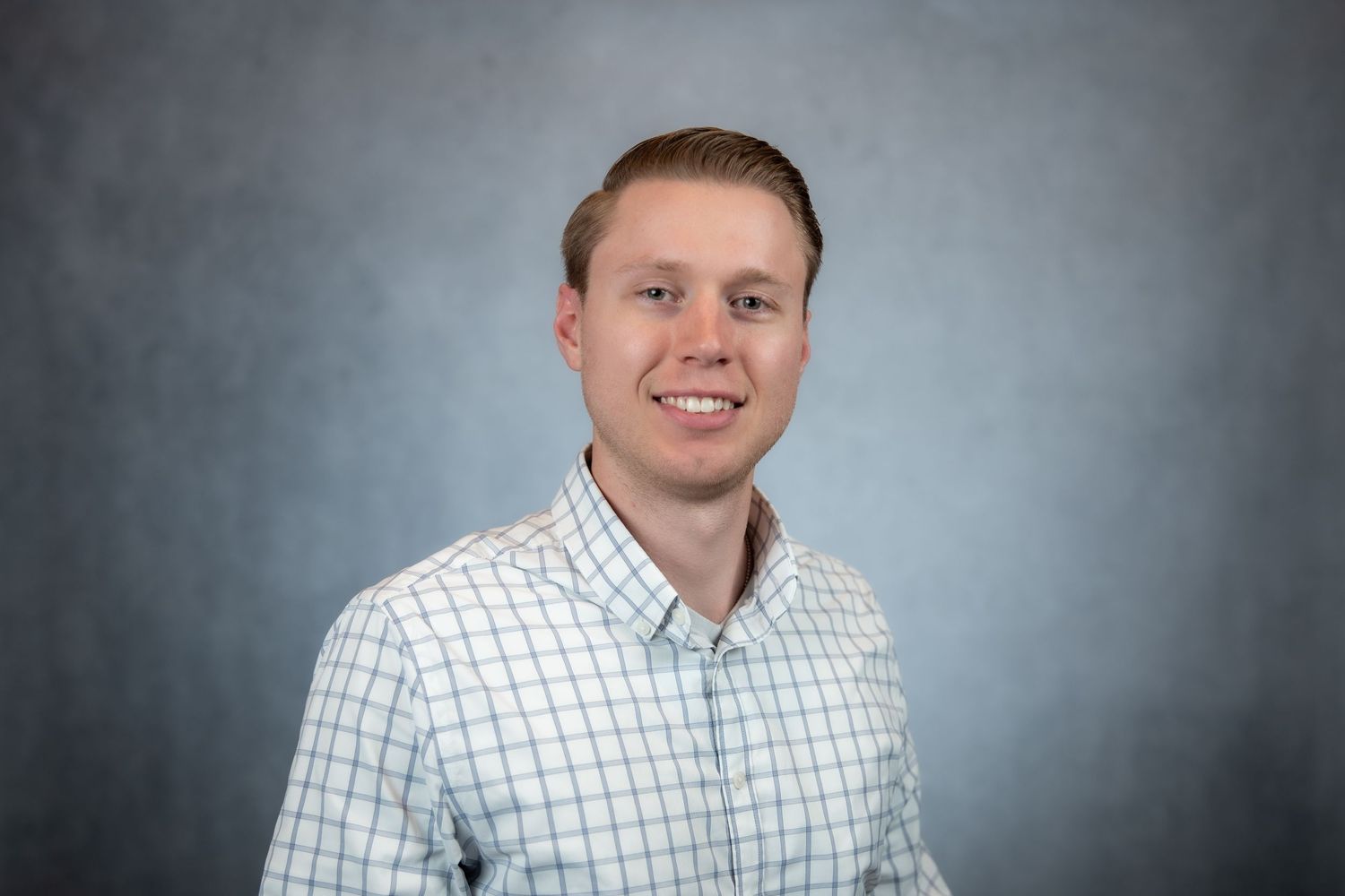 Smiling young man in light blue checkered shirt posing for professional headshot on gray background.