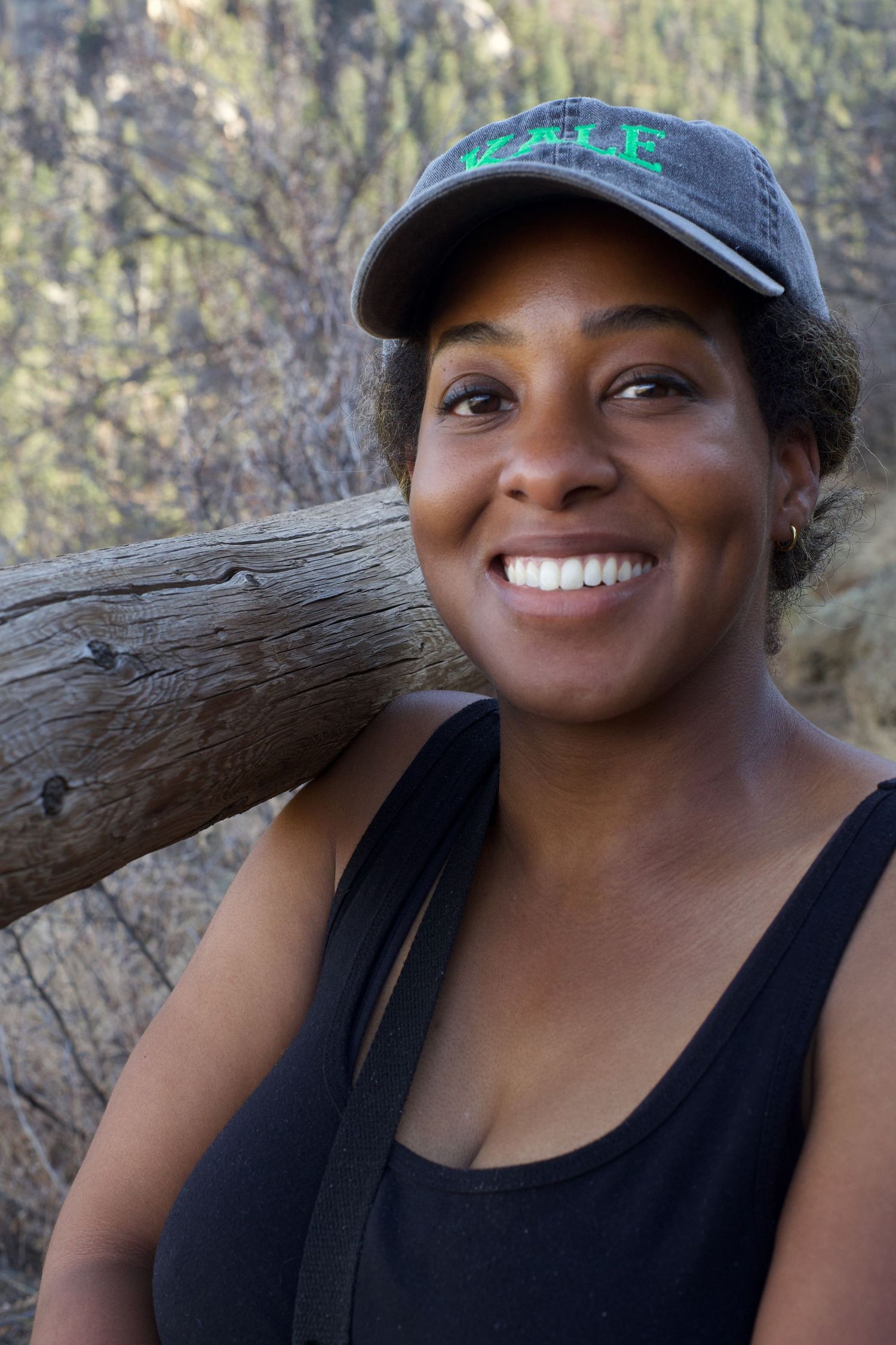 Smiling Black woman wearing a baseball cap and black tank top leaning against a log outdoors.