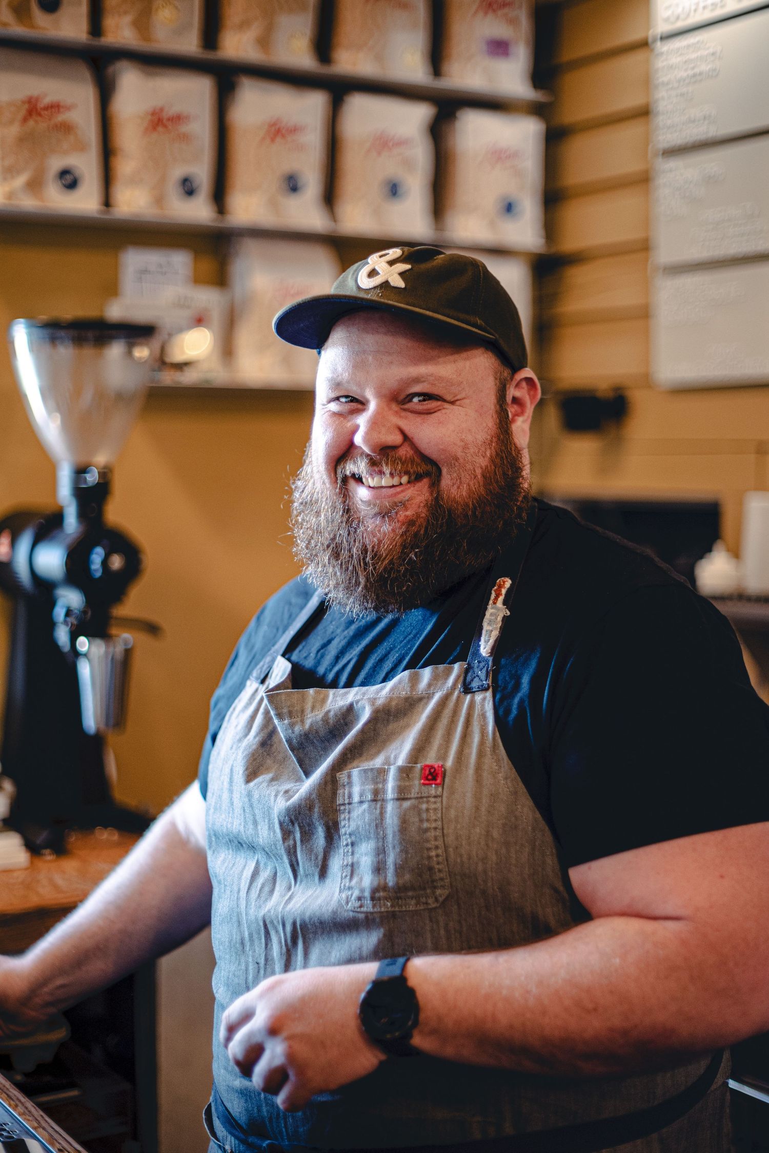 Smiling bearded barista in apron and cap standing behind coffee shop counter.