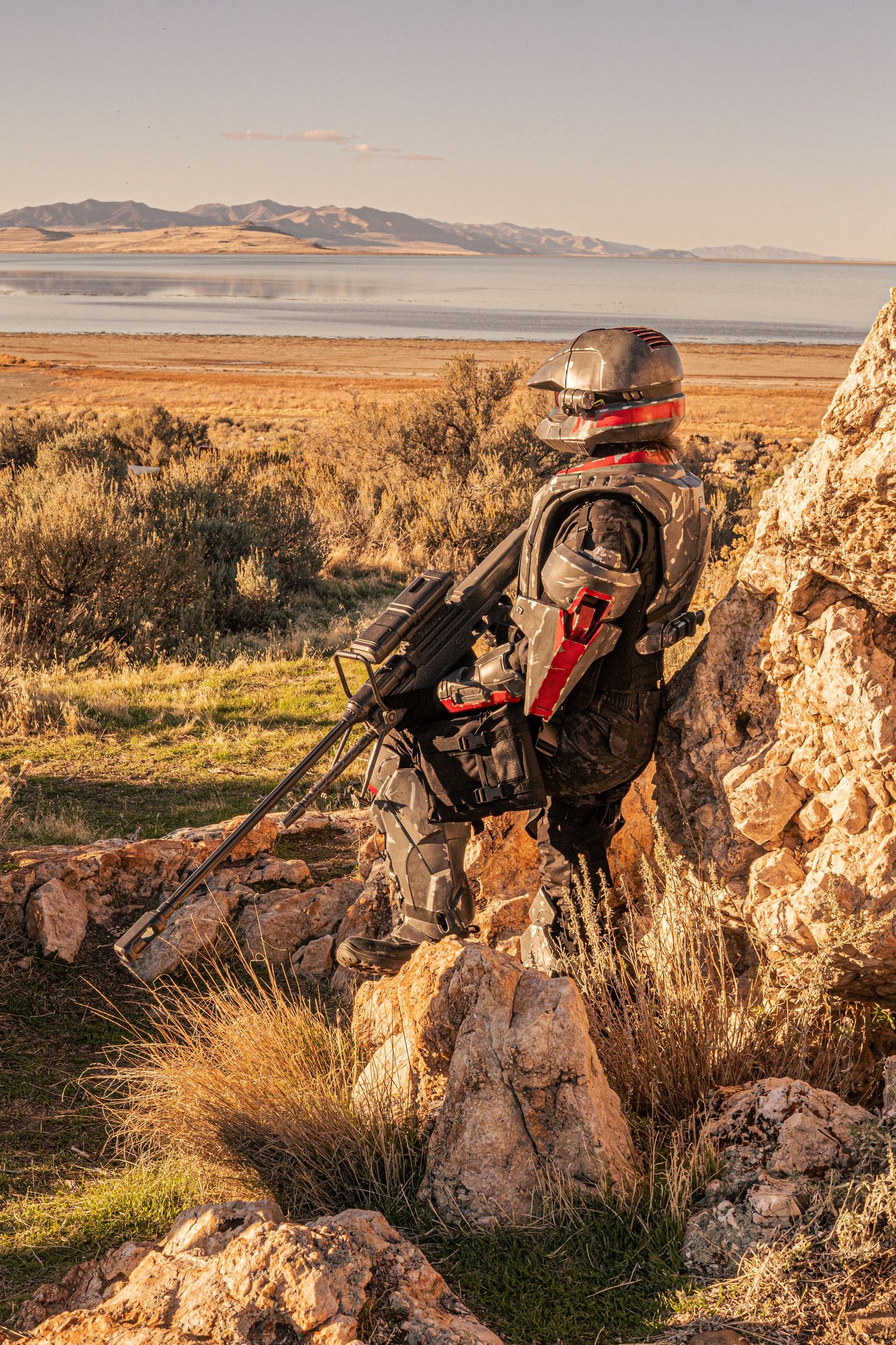 Armored cosplayer with jetpack stands on rocks overlooking a vast lake and mountain landscape at golden hour.