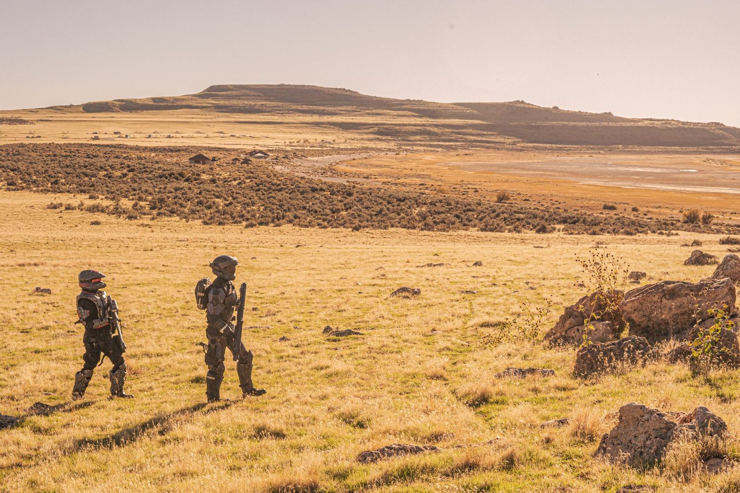 Two soldiers in futuristic armor suits patrol a vast golden grassland with rocky hills in the background.
