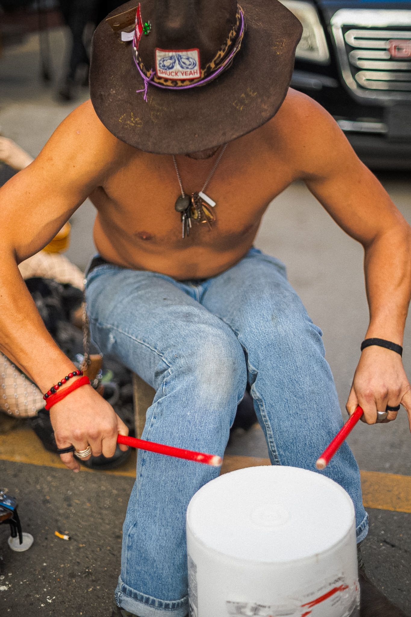 Shirtless street performer drumming on a white bucket with red sticks, wearing a cowboy hat and jeans outdoors.