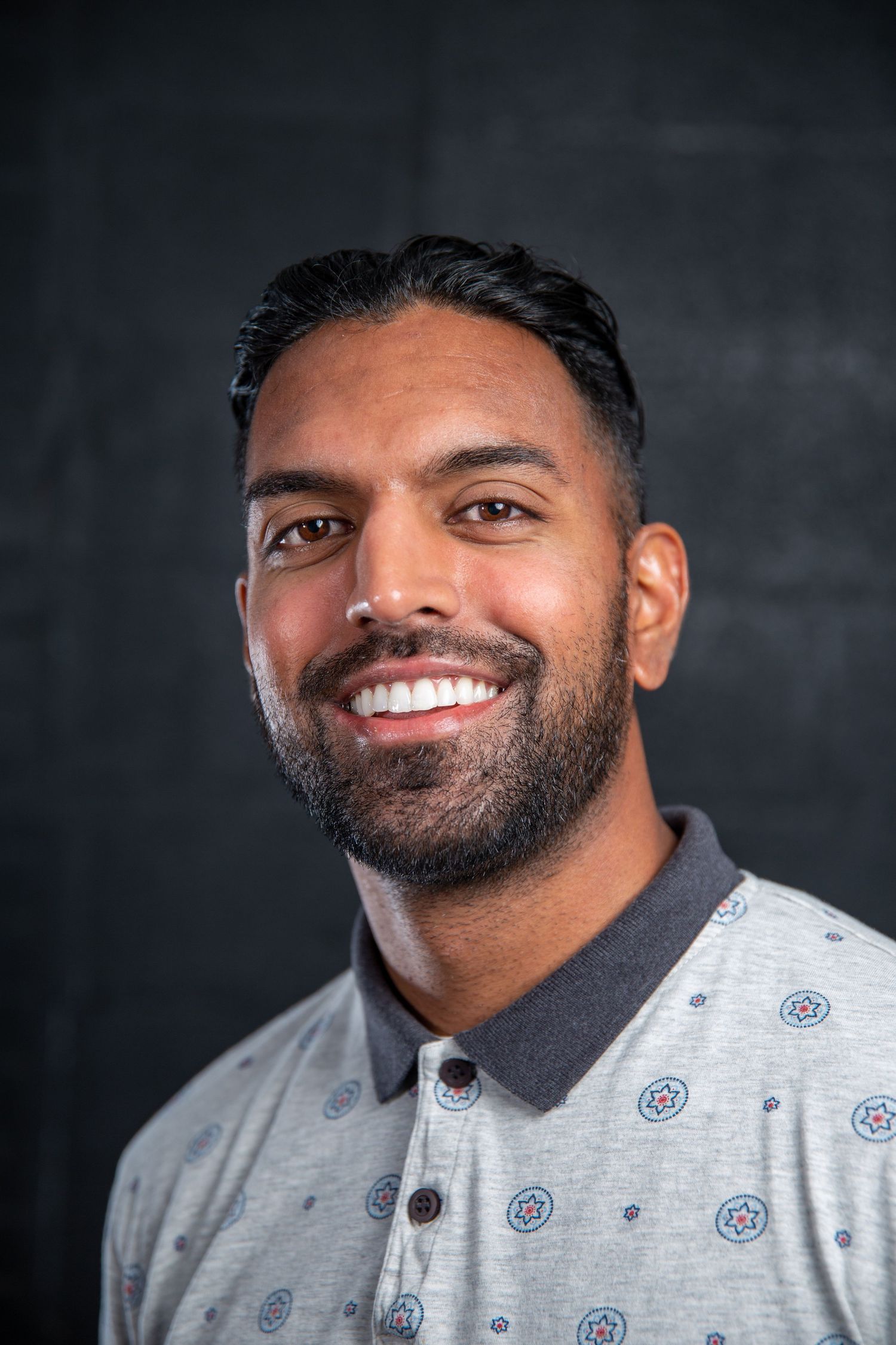 Smiling bearded man in a grey patterned polo shirt poses for a professional portrait photo.