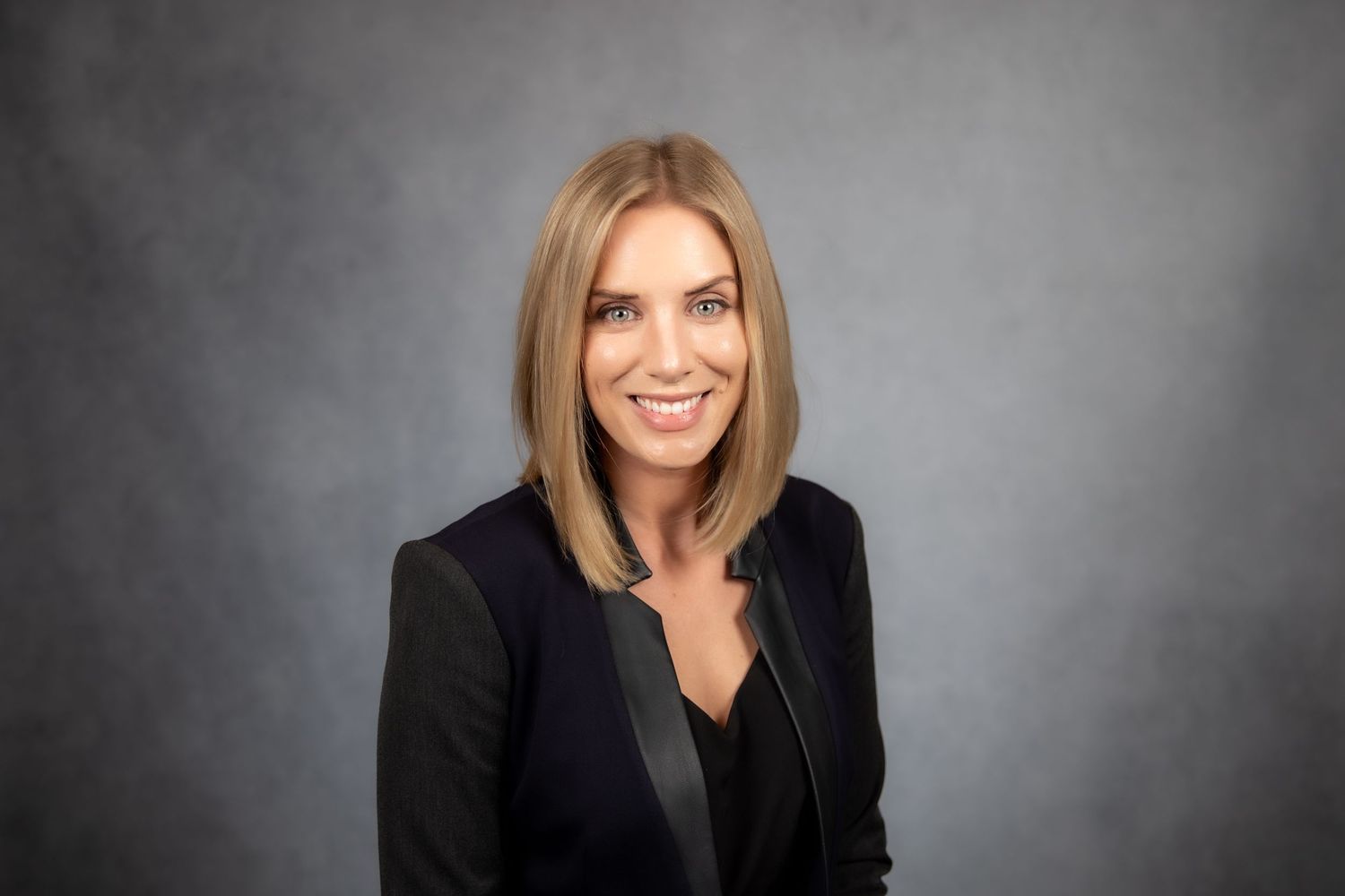 Blonde woman smiling in a black blazer posing for a professional headshot against a gray background.