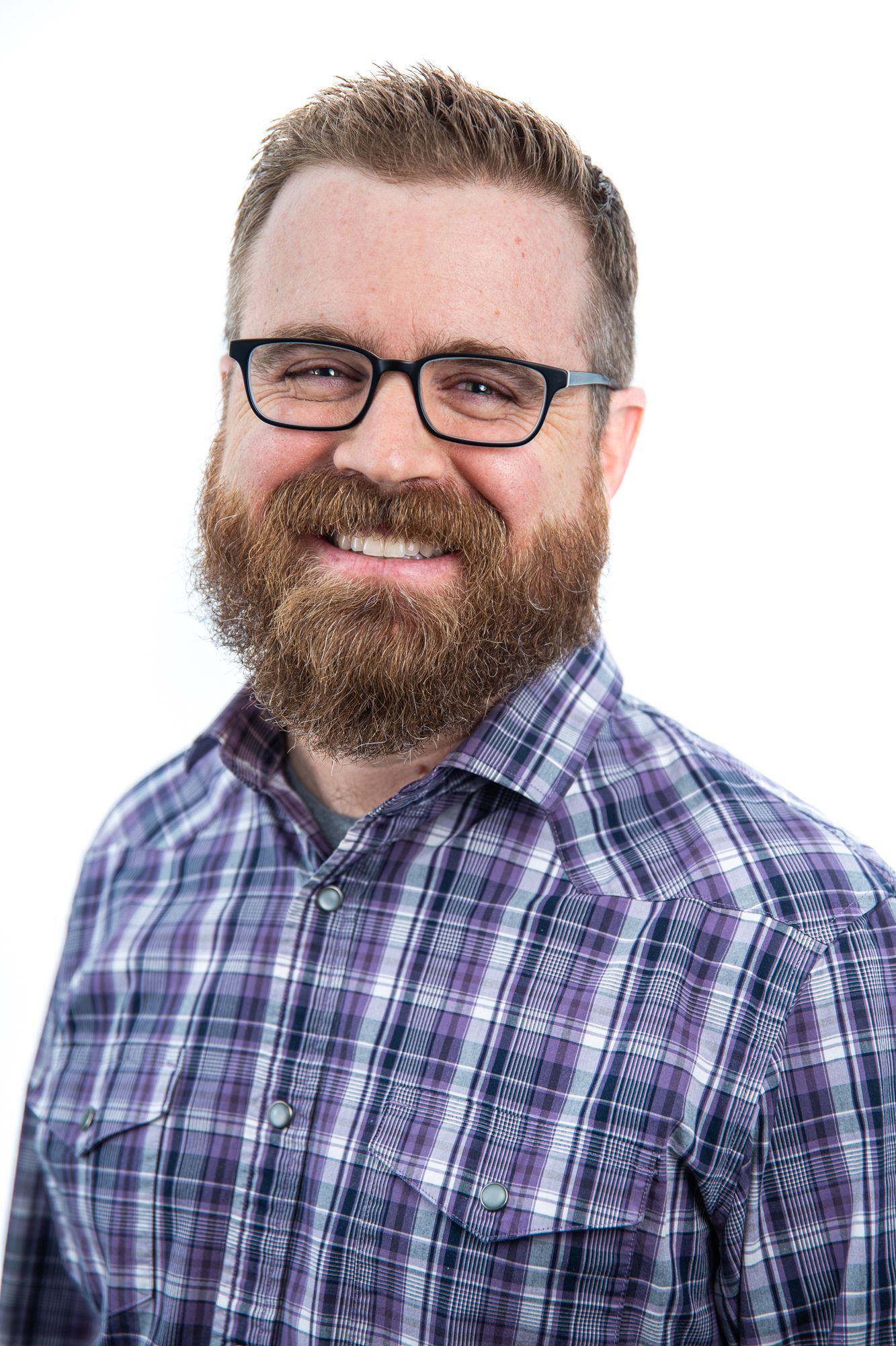 Bearded man with glasses smiling in a plaid shirt against a white background.