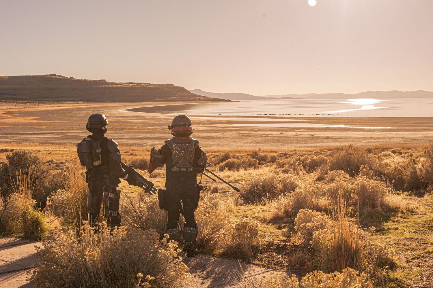 Two sci-fi armored cosplayers overlooking vast desert lake at golden sunset.