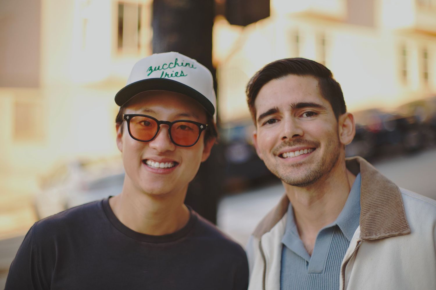 Two smiling young men posing together outdoors in warm golden light, one wearing sunglasses and a cap.