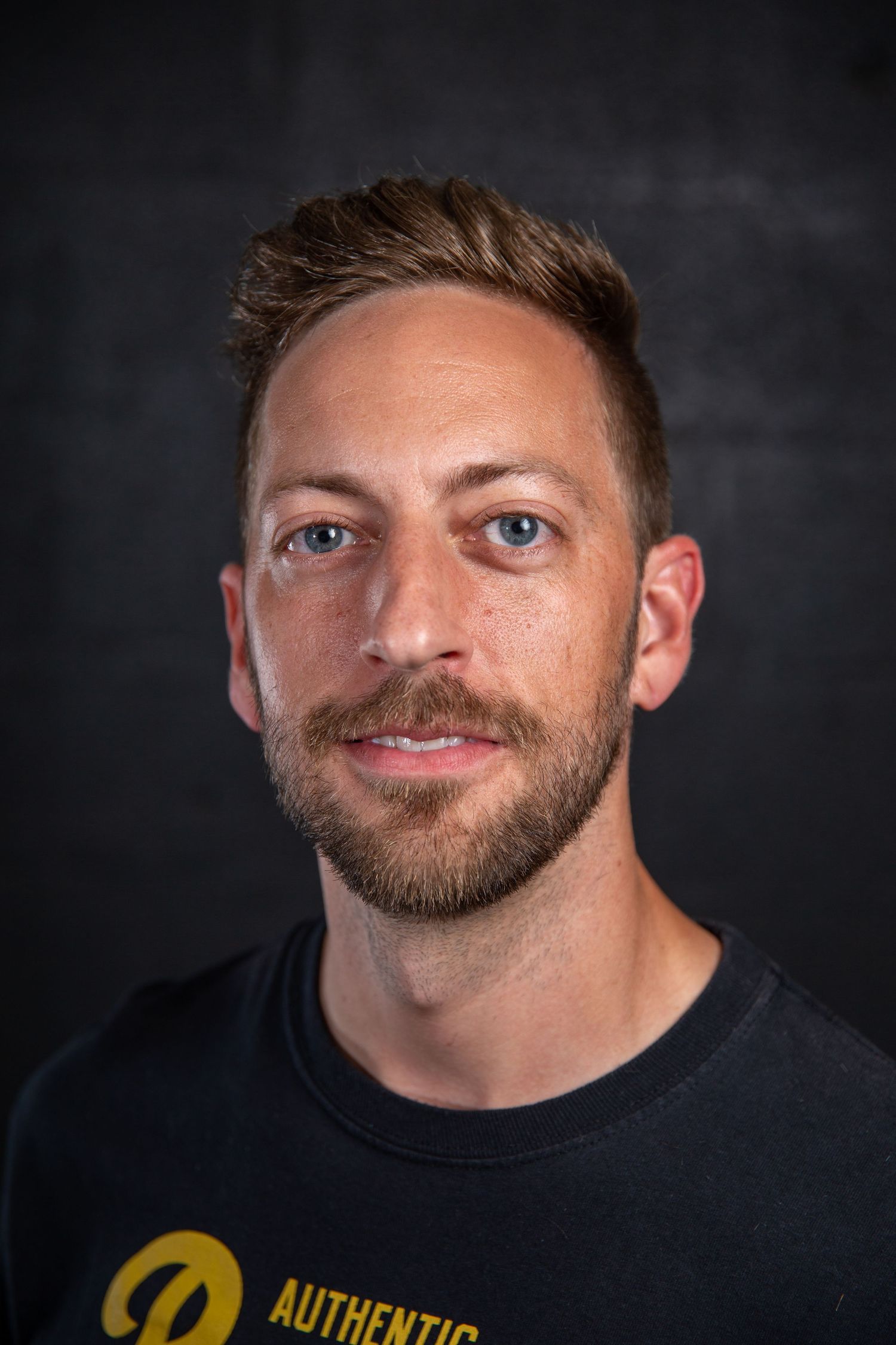 Caucasian man with short hair and beard wearing a black Authentic brand t-shirt against dark background.