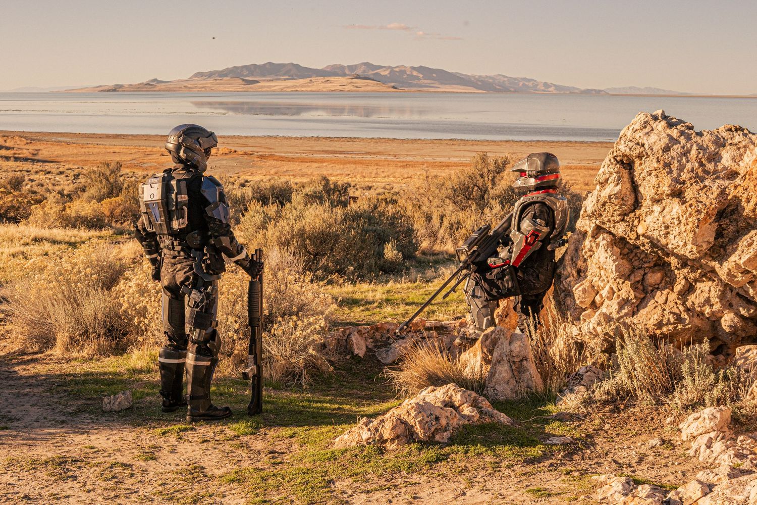 Two cosplayers in futuristic combat armor suits posing near rocky terrain with a lake and mountains behind.