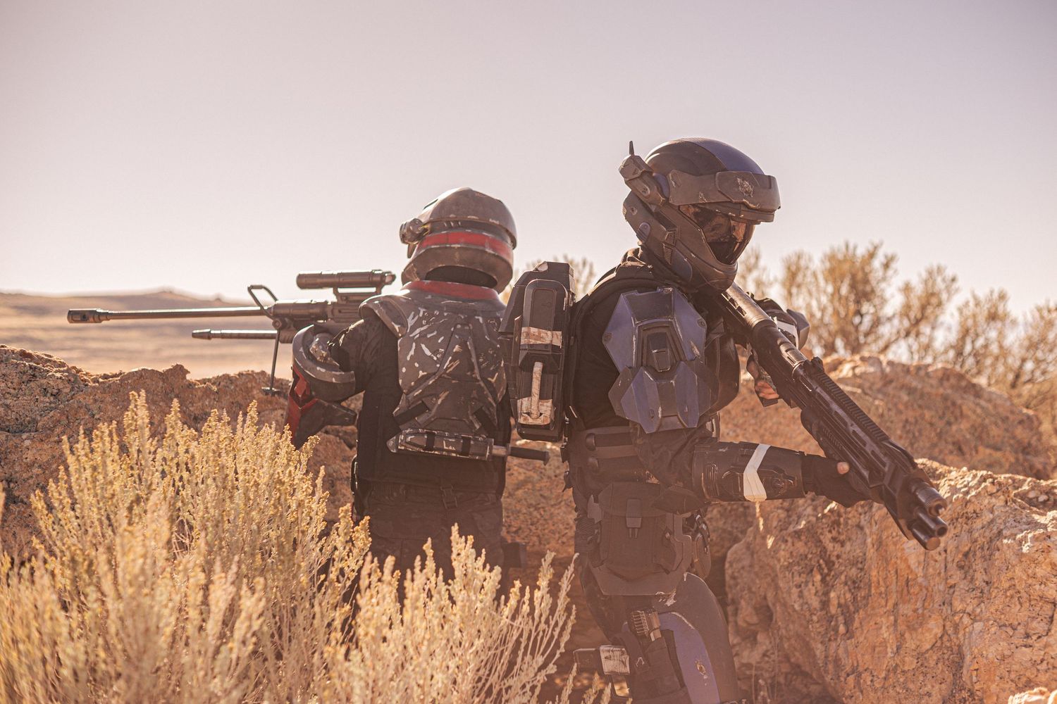 Two armored cosplayers with futuristic gear and weapons survey a desert landscape with shrubs and a lake in the background.
