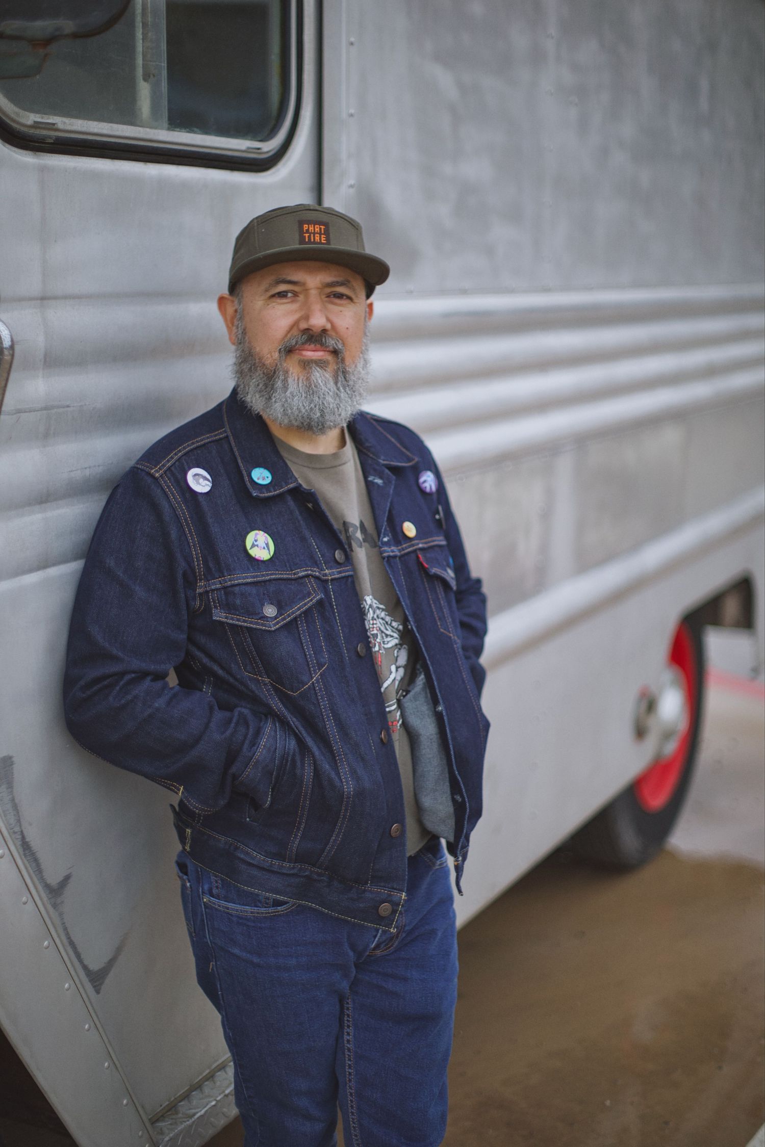 Bearded man in denim jacket with pins standing beside a silver trailer outdoors.