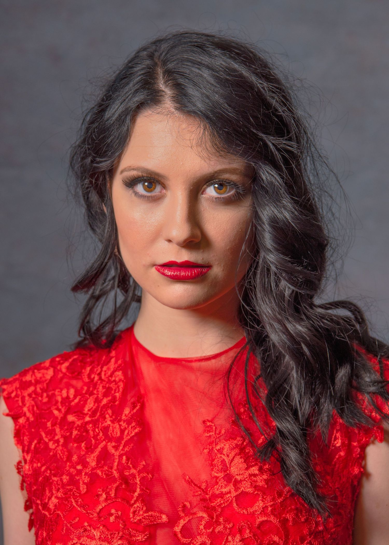 Young woman with dark wavy hair and red lips wearing a red lace dress against a gray background.