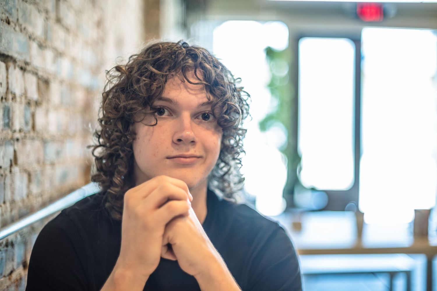Young man with curly hair sitting thoughtfully with hands clasped near a brick wall in a cafe.