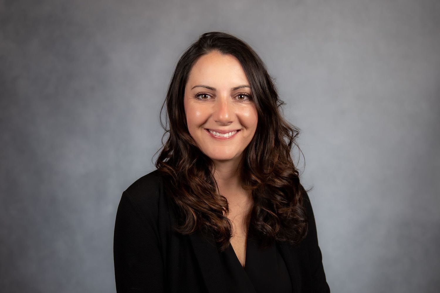 Professional headshot of a smiling woman with curly brown hair wearing a black top against a gray background.