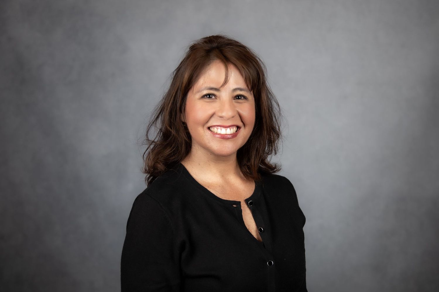 Friendly woman with shoulder-length brown hair wearing a black top, professional headshot on gray background.
