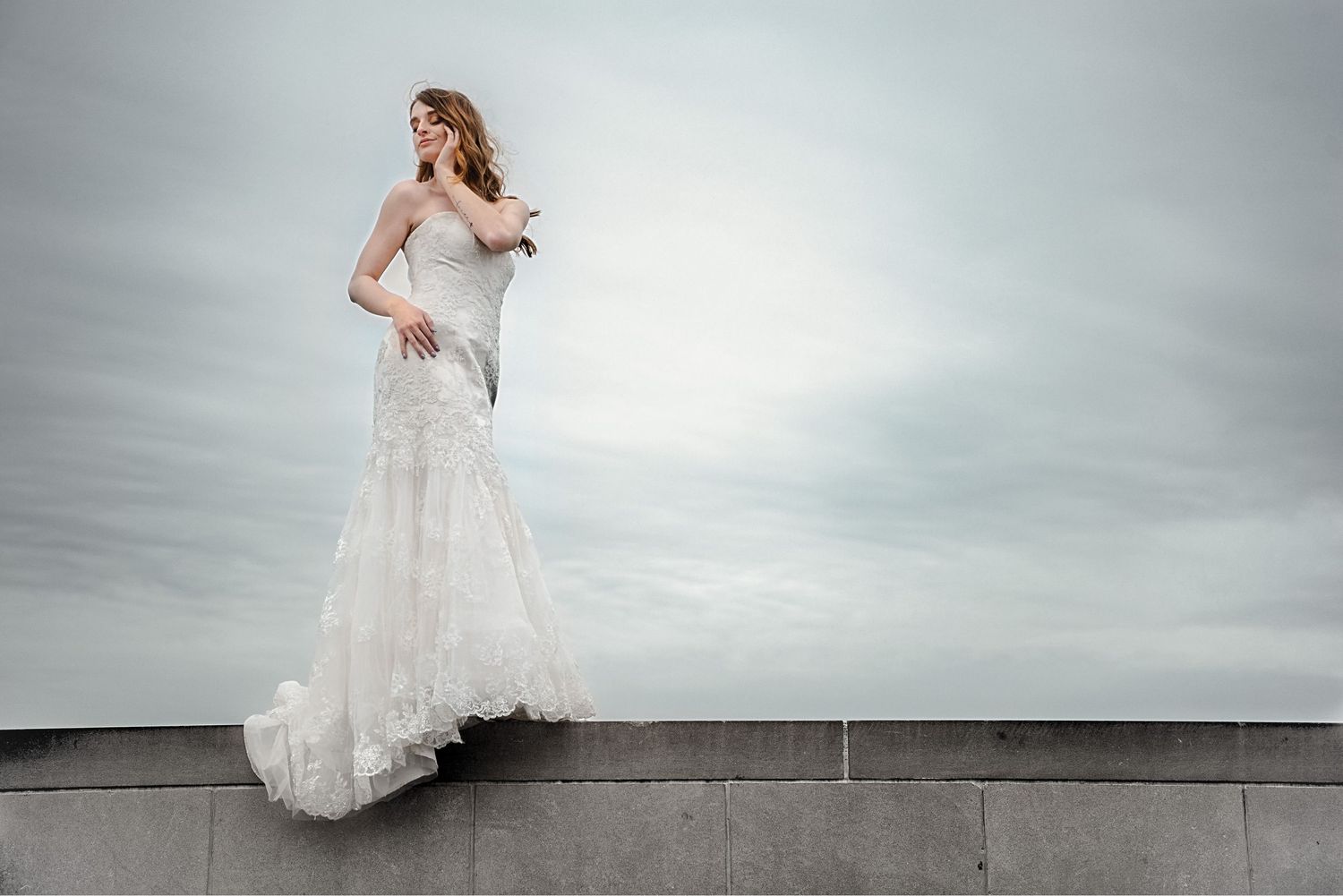 Elegant bride in a white lace mermaid wedding gown standing on a stone wall against a cloudy sky.
