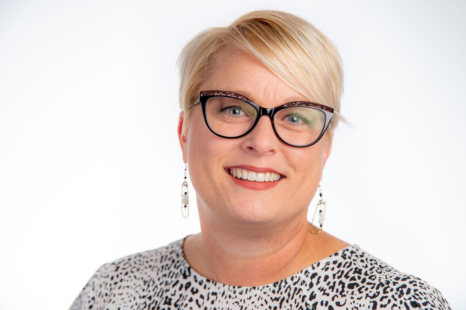 Smiling blonde woman wearing glasses and patterned top with dangling earrings on white background.